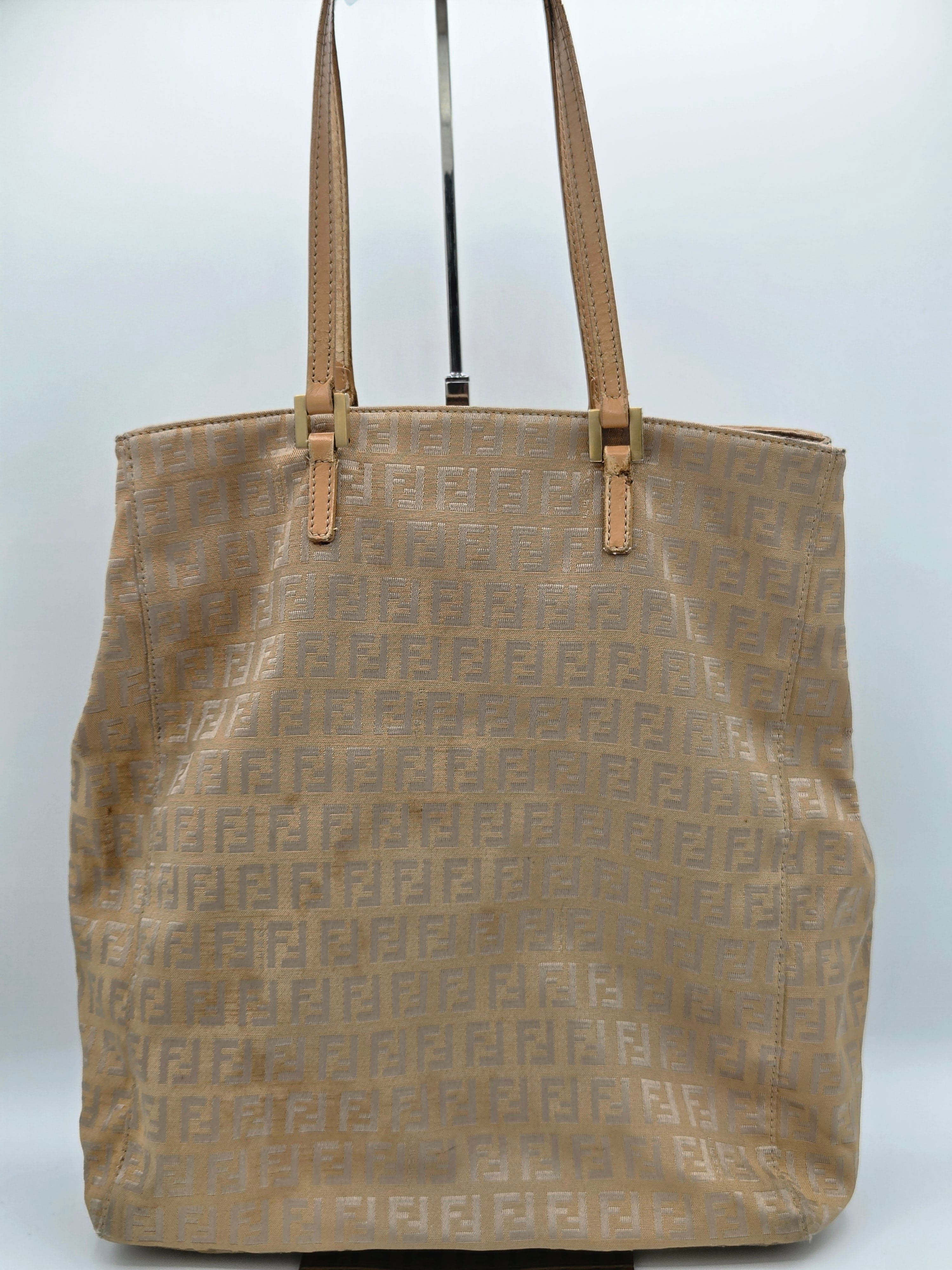 A beige Fendi tote bag with an all-over embossed FF monogram pattern and tan leather shoulder straps, shown hanging against a plain light gray background.