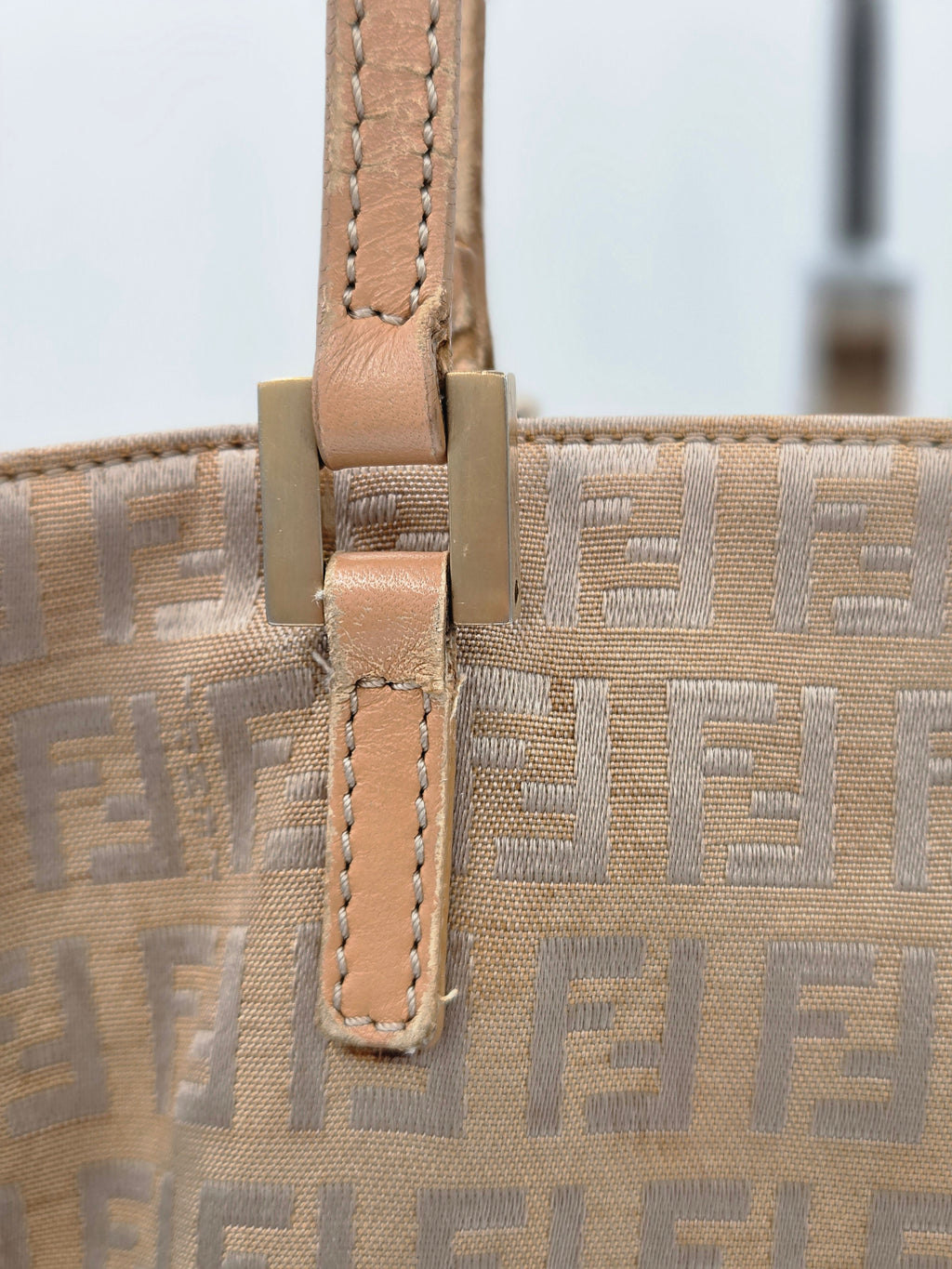 Close-up of a beige Fendi tote bag featuring the signature FF monogram canvas and a tan leather strap with brass-tone hardware.