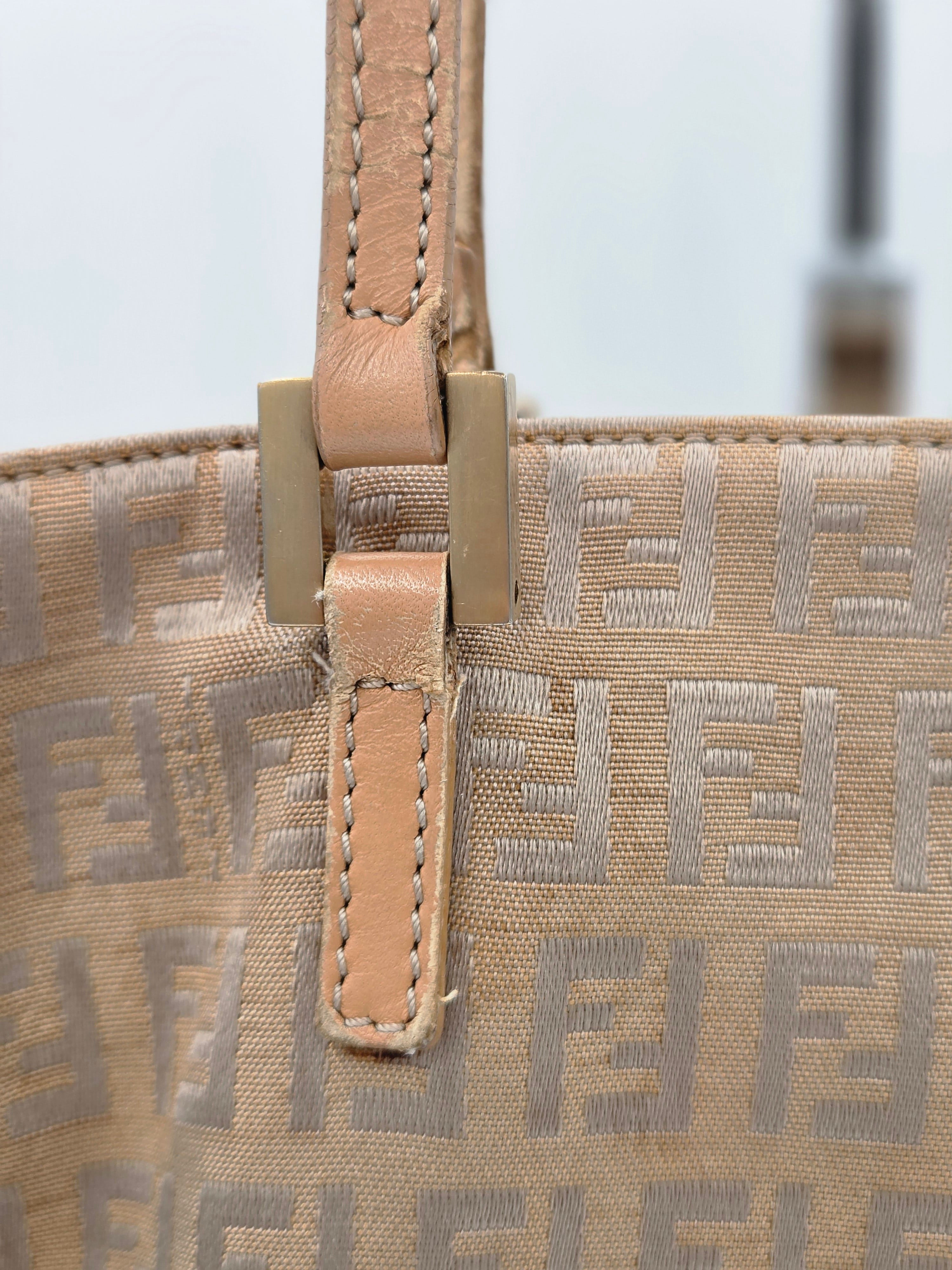 Close-up of a beige Fendi tote bag featuring the signature FF monogram canvas and a tan leather strap with brass-tone hardware.