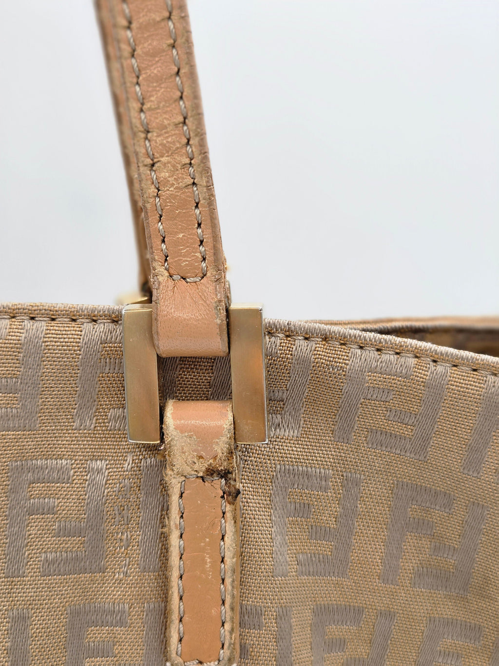 Close-up of a beige Fendi handbag featuring the signature Zucca monogram canvas and a tan leather strap secured with a gold-tone metal buckle.