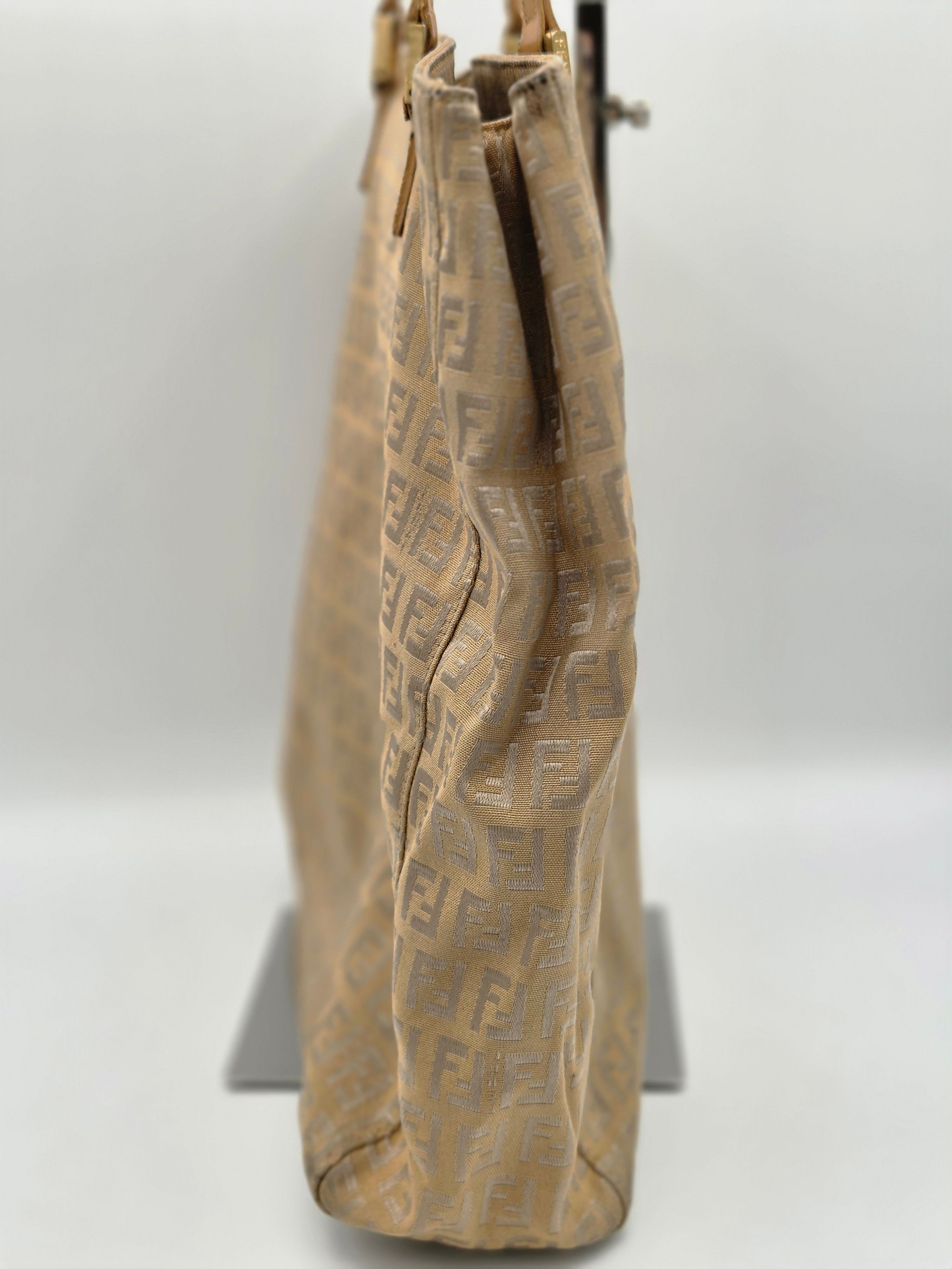 A beige Fendi monogram canvas shoulder bag with a gold-tone metal clasp, shown hanging against a plain white wall.