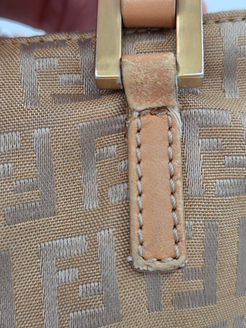 Close-up of a beige woven monogram fabric with a tan leather tab and gold-tone metal hardware, showing the texture and stitching detail.
