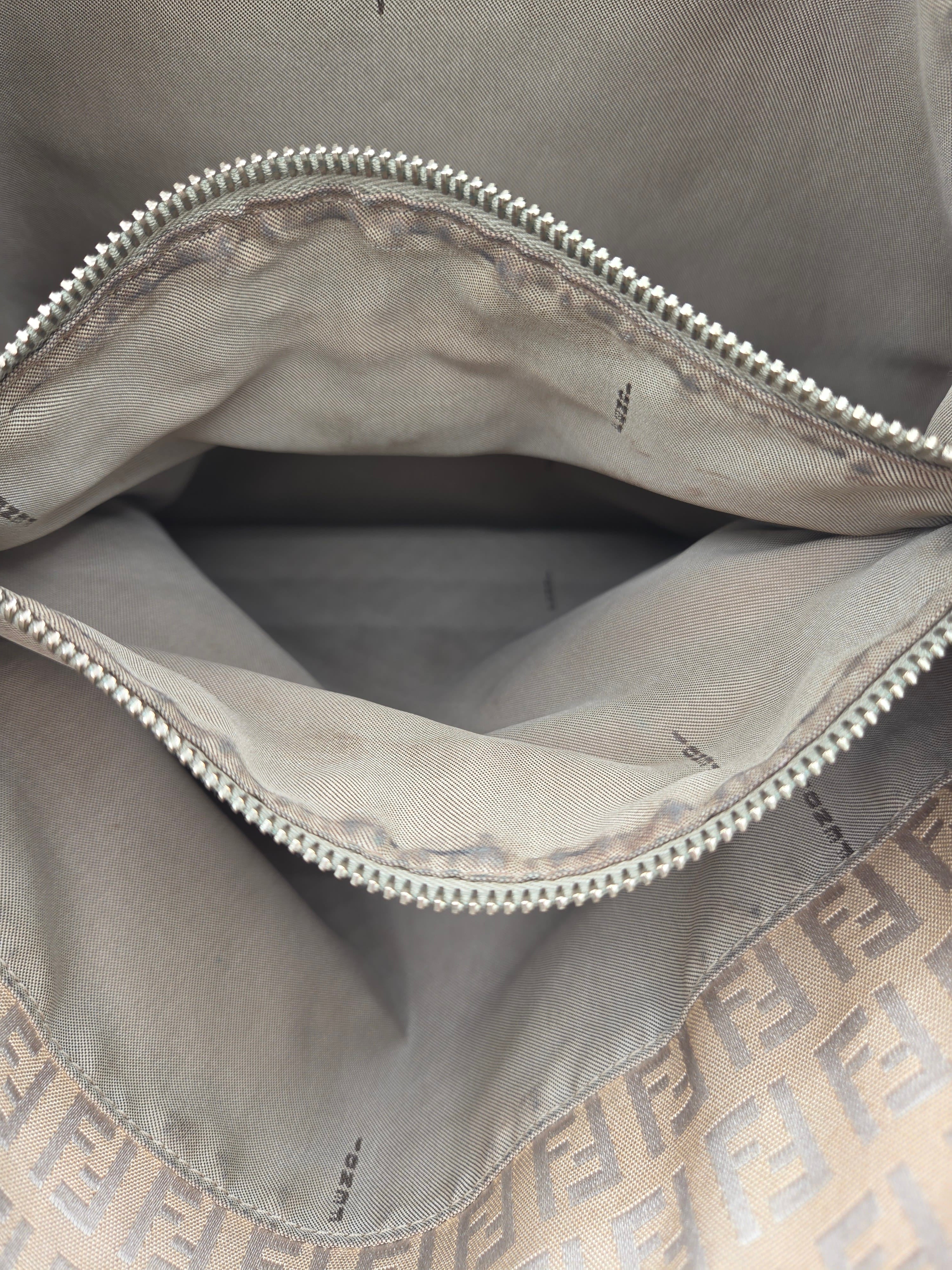 Close-up of the interior of a beige handbag or clutch, showing a silver-tone zipper, beige interior lining, and a tan monogram border with an FF pattern at the bottom.
