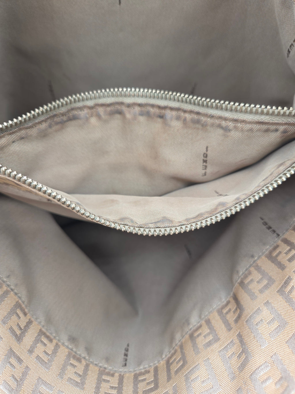 Close-up of the interior of a beige Fendi bag with a silver zipper and the signature Zucca monogram pattern visible along the bottom edge.