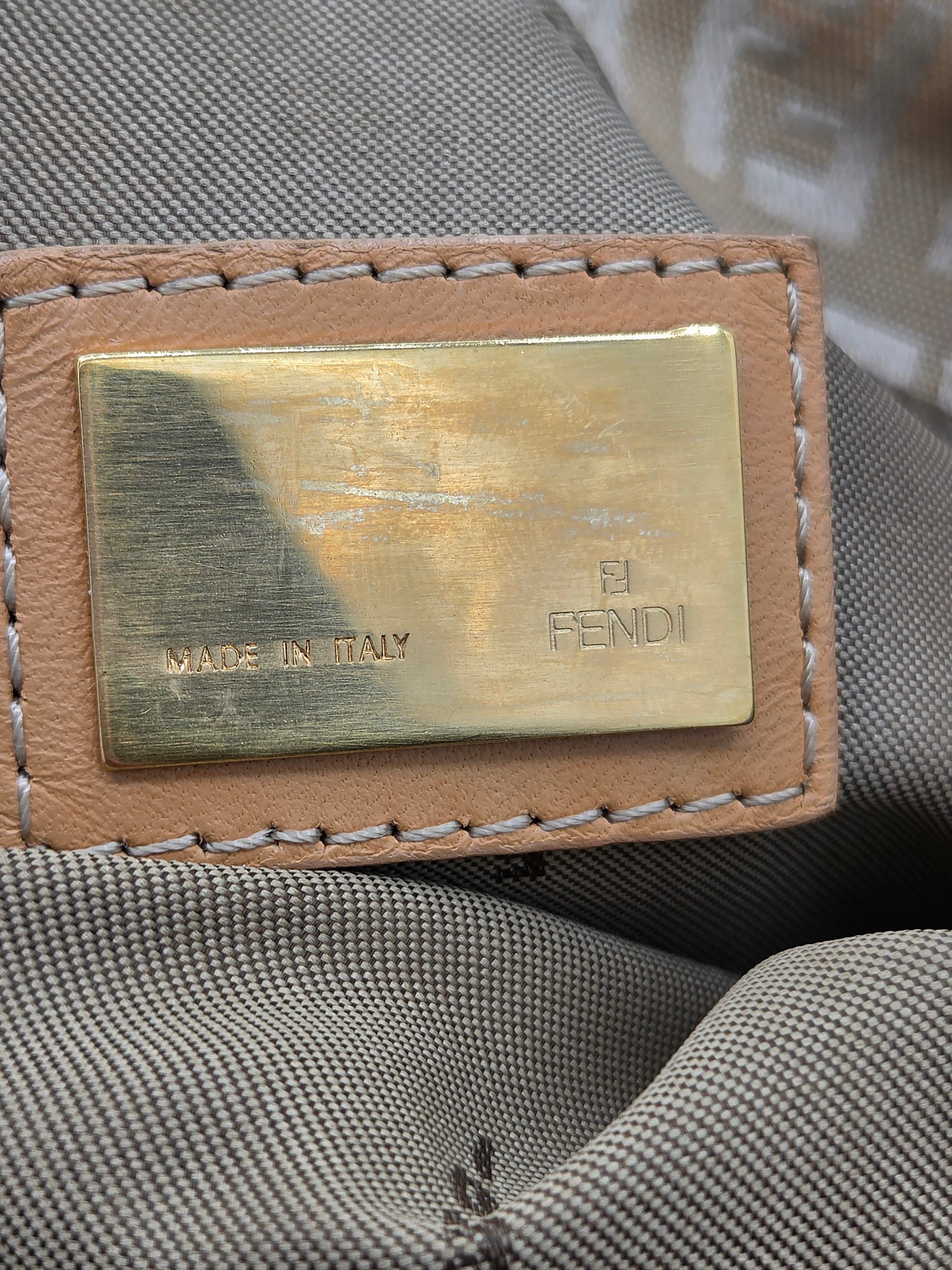 Close-up of a Fendi metal plaque on a light brown leather patch, attached to a gray woven fabric.