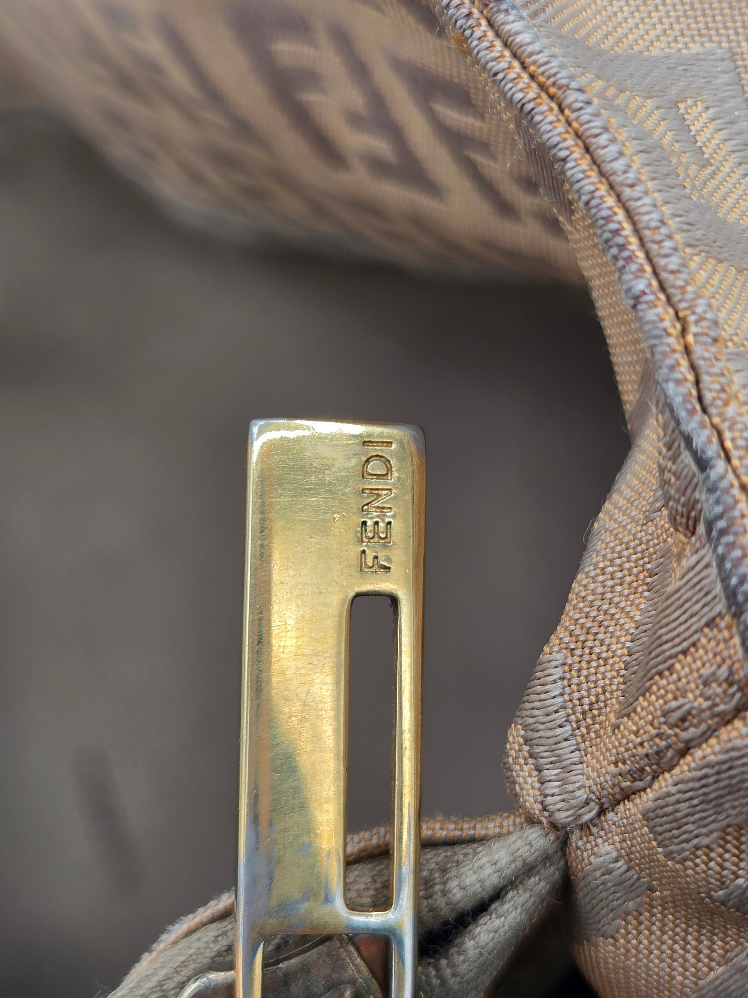 Close-up of a gold-toned metal zipper pull engraved with 'FENDI', attached to a beige and brown patterned fabric strap.