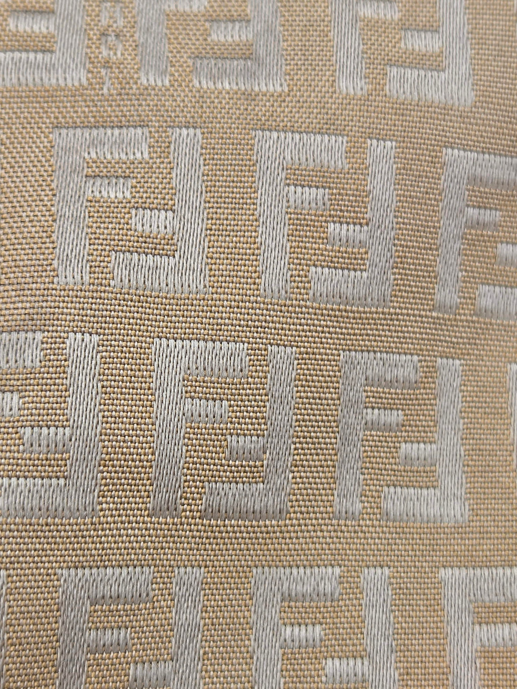 Close-up of a woven fabric with a repeating geometric monogram pattern in light gray on a warm beige background.