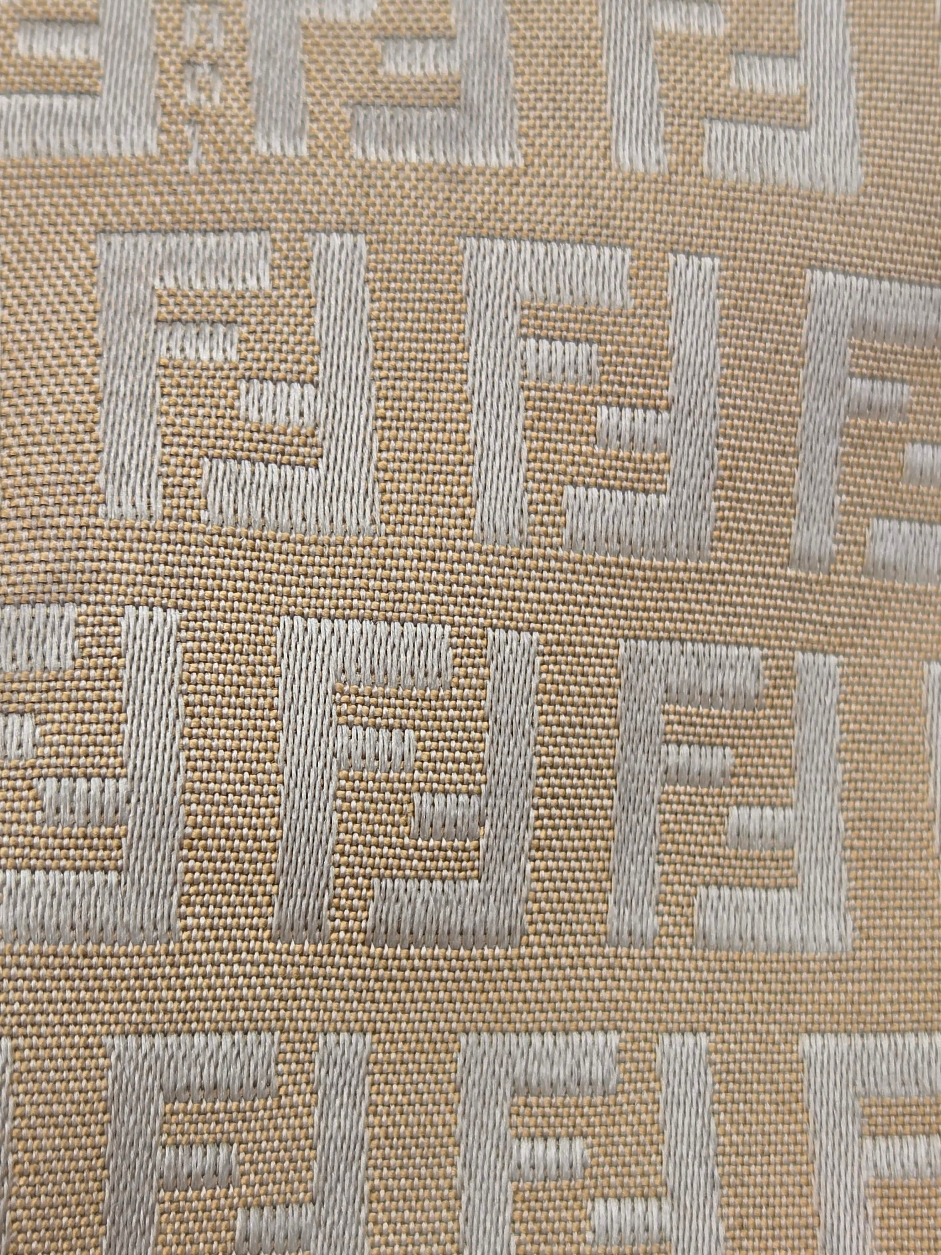 Close-up of a woven fabric with a repeating geometric monogram pattern in light gray on a warm beige background.