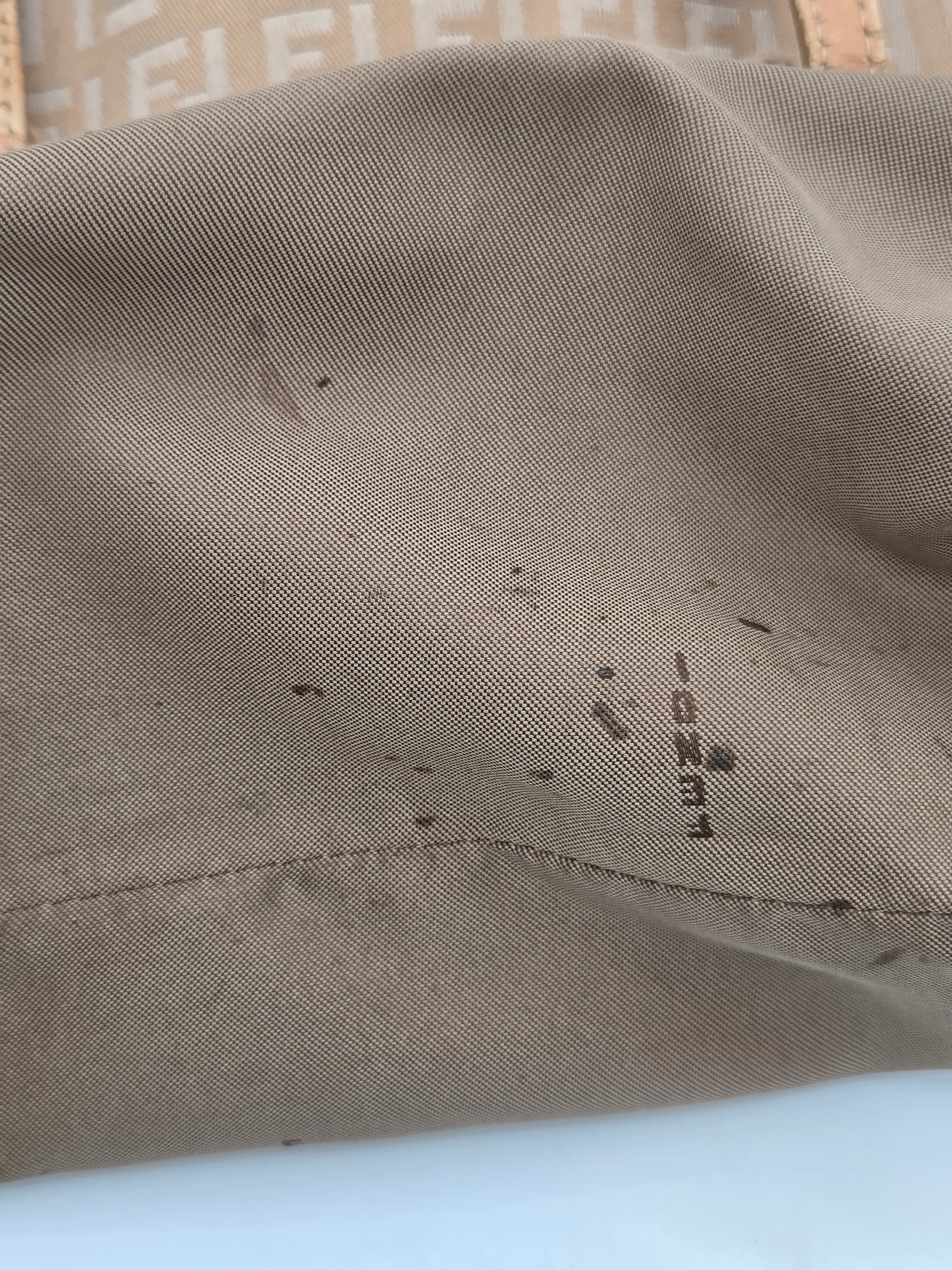 Close-up of a beige or taupe woven fabric surface with a small dark printed marking and scattered small dark specks, photographed against a light neutral background.