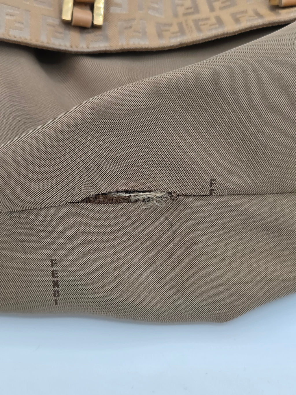 Close-up of a taupe brown tweed garment, showing a vertical seam with frayed thread and small black embroidered text near the lower right, alongside a patterned beige strap on the left.