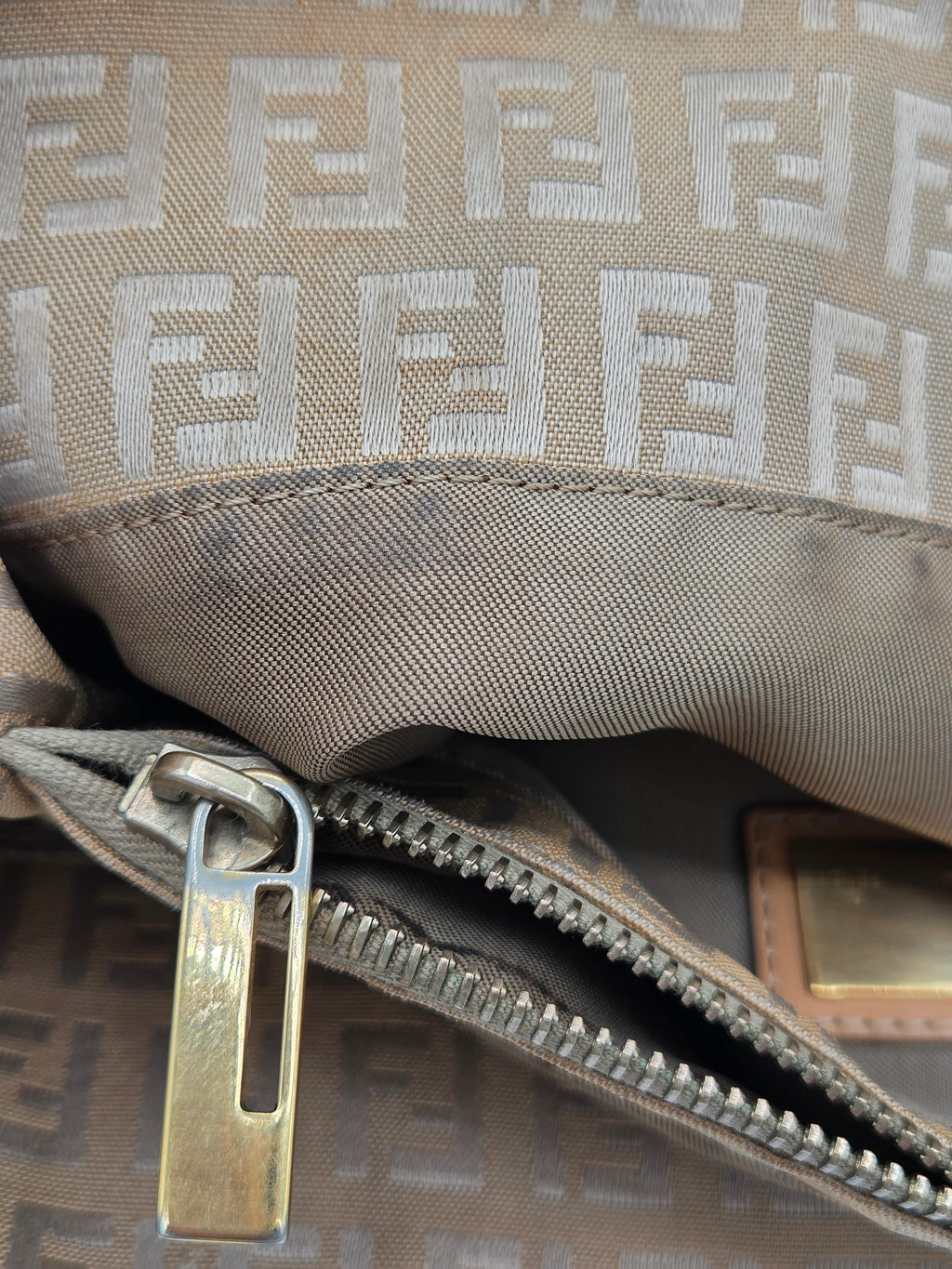 Close-up of a Fendi bag in brown and beige monogram canvas, showing the iconic FF logo pattern, a silver zipper with a brass pull, and a small gold-tone brand plaque.