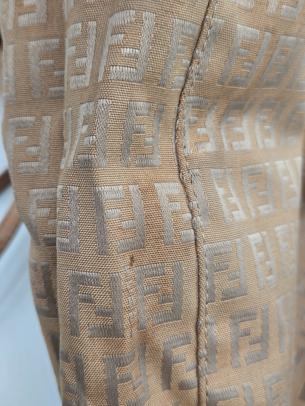 Close-up of a beige woven textile with a repeating monogram pattern in a light gray tone, showing fabric texture and stitching.