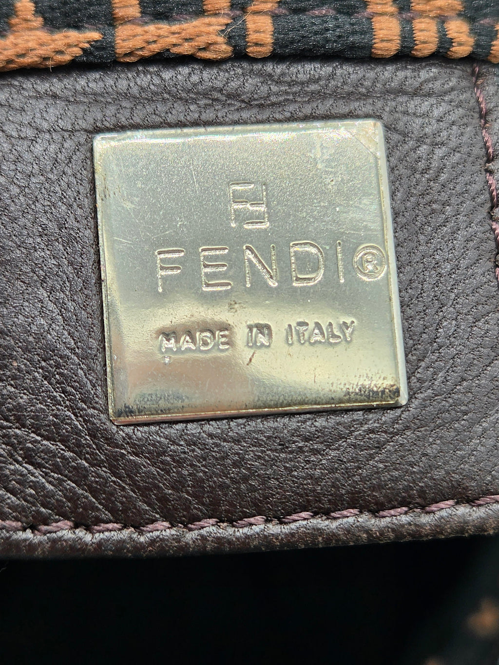 Close-up of a Fendi leather interior label with a metallic gold-toned plaque reading FENDI and MADE IN ITALY, attached to dark brown textured leather.