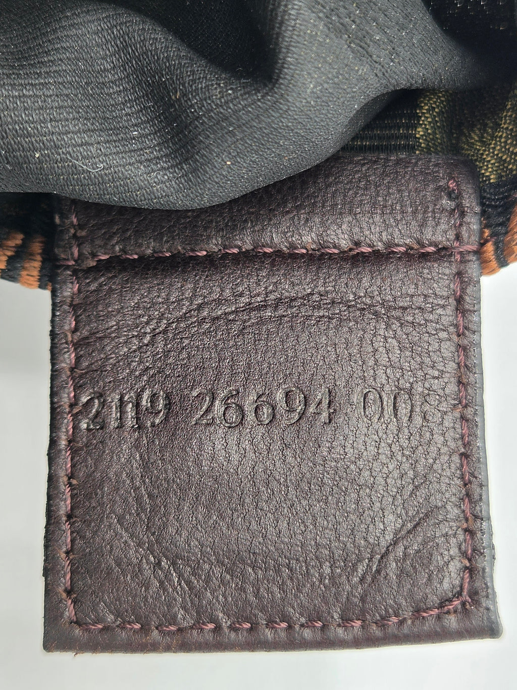 Close-up of a dark brown leather patch with embossed serial numbers '2119 26694 on' and matching stitching, with dark gray fabric and a plaid pattern visible above.