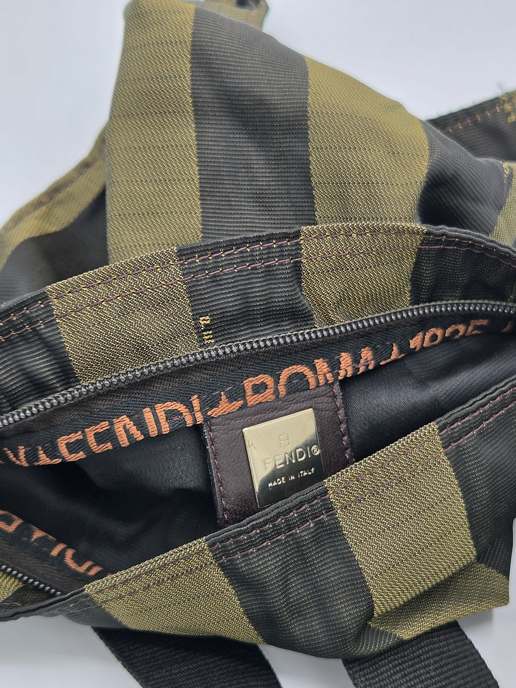 Close-up of a checkered bag interior with olive green and black stripes, a black zipper track with gold-tone lettering, and a small gold brand patch.