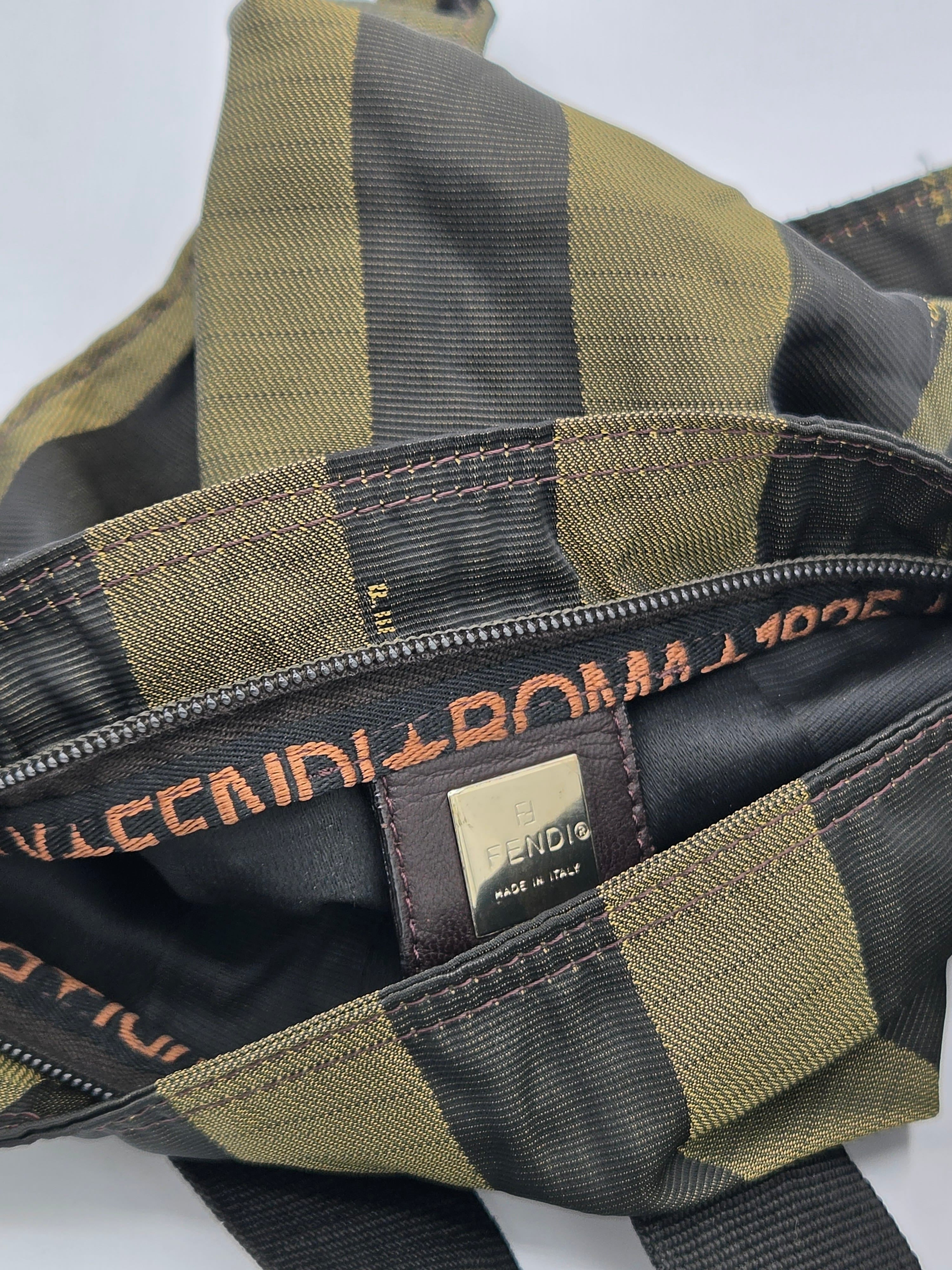 Close-up of a checkered bag interior with olive green and black stripes, a black zipper track with gold-tone lettering, and a small gold brand patch.