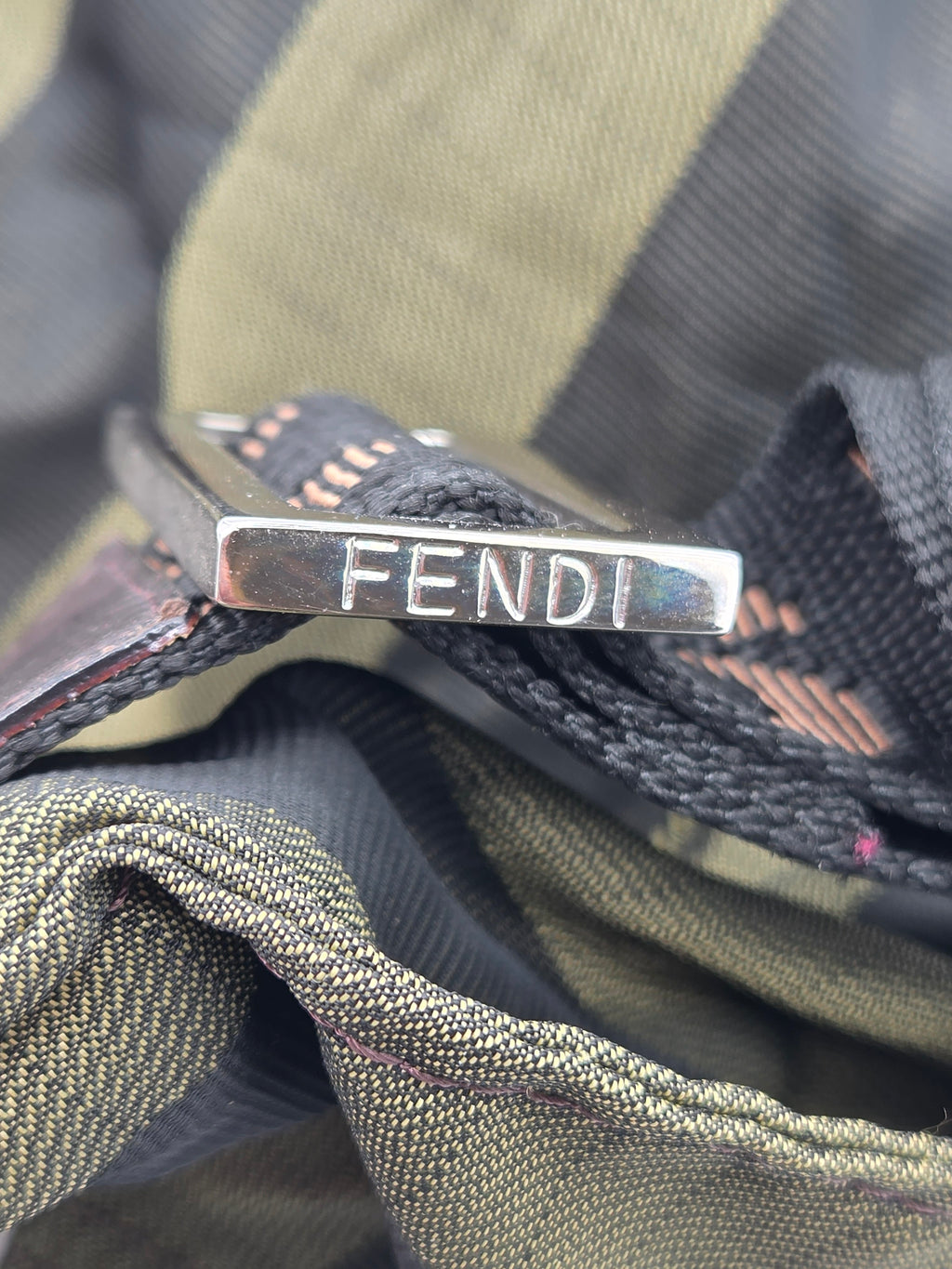 A close-up of a silver metal Fendi logo bar attached to a black woven strap, resting on olive green and black striped fabric.