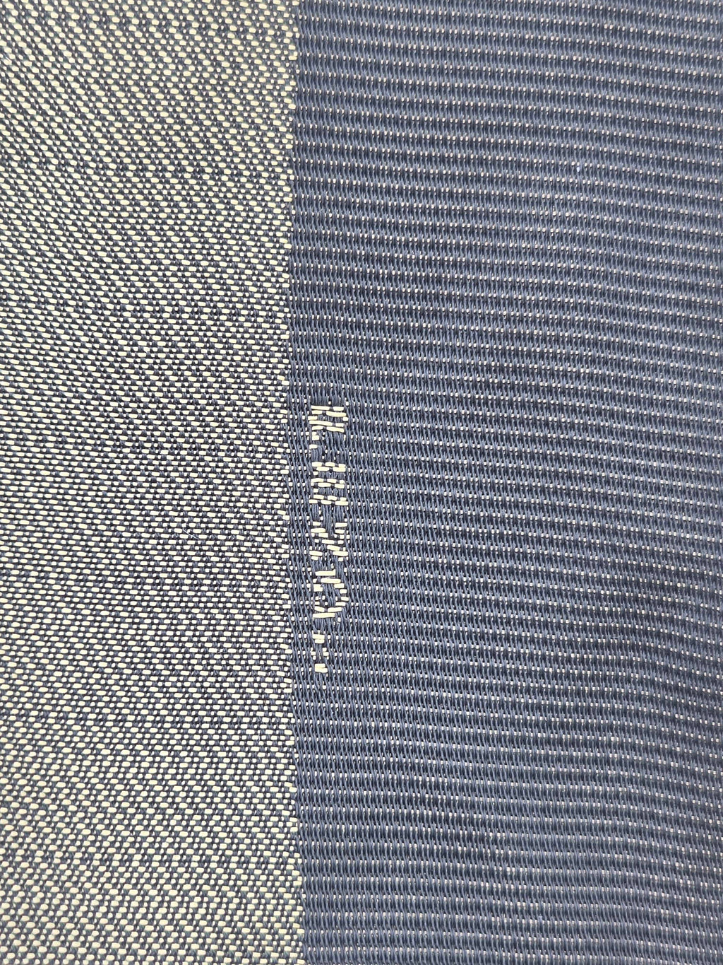 Close-up of a woven textile with a vertical color-blocked pattern, featuring a light beige/cream textured section on the left and a slate blue textured section on the right. The brand name 'Helly Hansen' is woven vertically in white between the two sections.