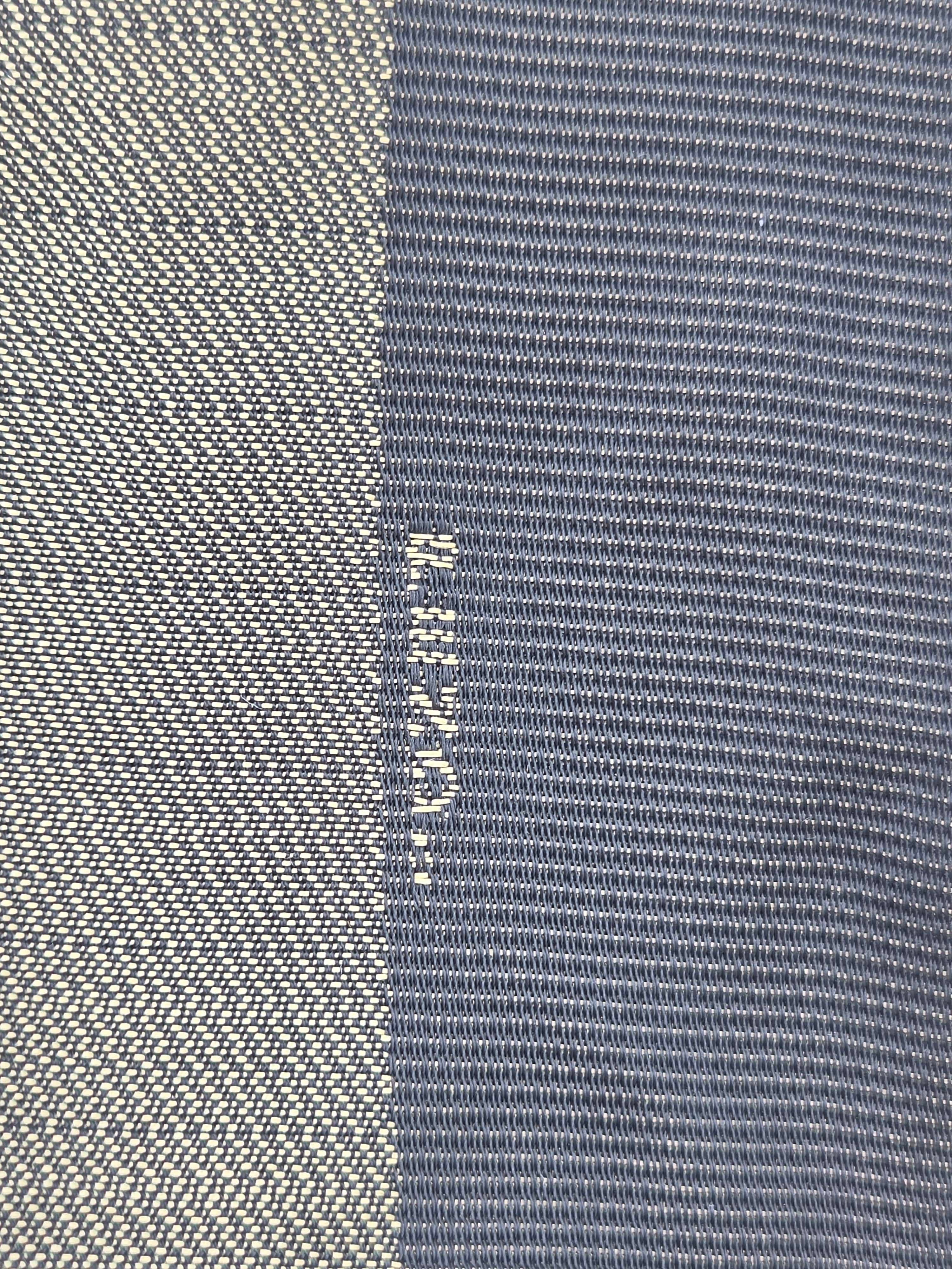 Close-up of a woven textile with a vertical color-blocked pattern, featuring a light beige/cream textured section on the left and a slate blue textured section on the right. The brand name 'Helly Hansen' is woven vertically in white between the two sections.