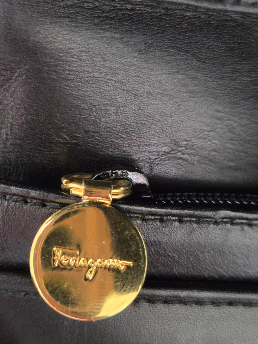 Close-up of a black leather zipper pull featuring a round polished gold-tone metal clasp with the Ferragamo logo engraved on it.