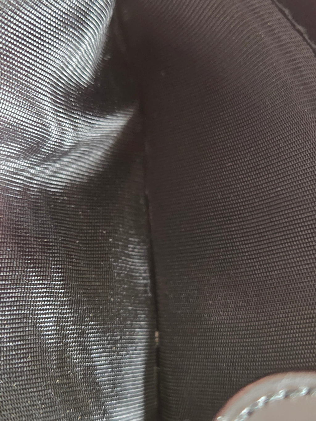 Close-up of a dark gray or black textured fabric surface with a small portion of a light-colored stitched circular detail visible in the upper right corner.