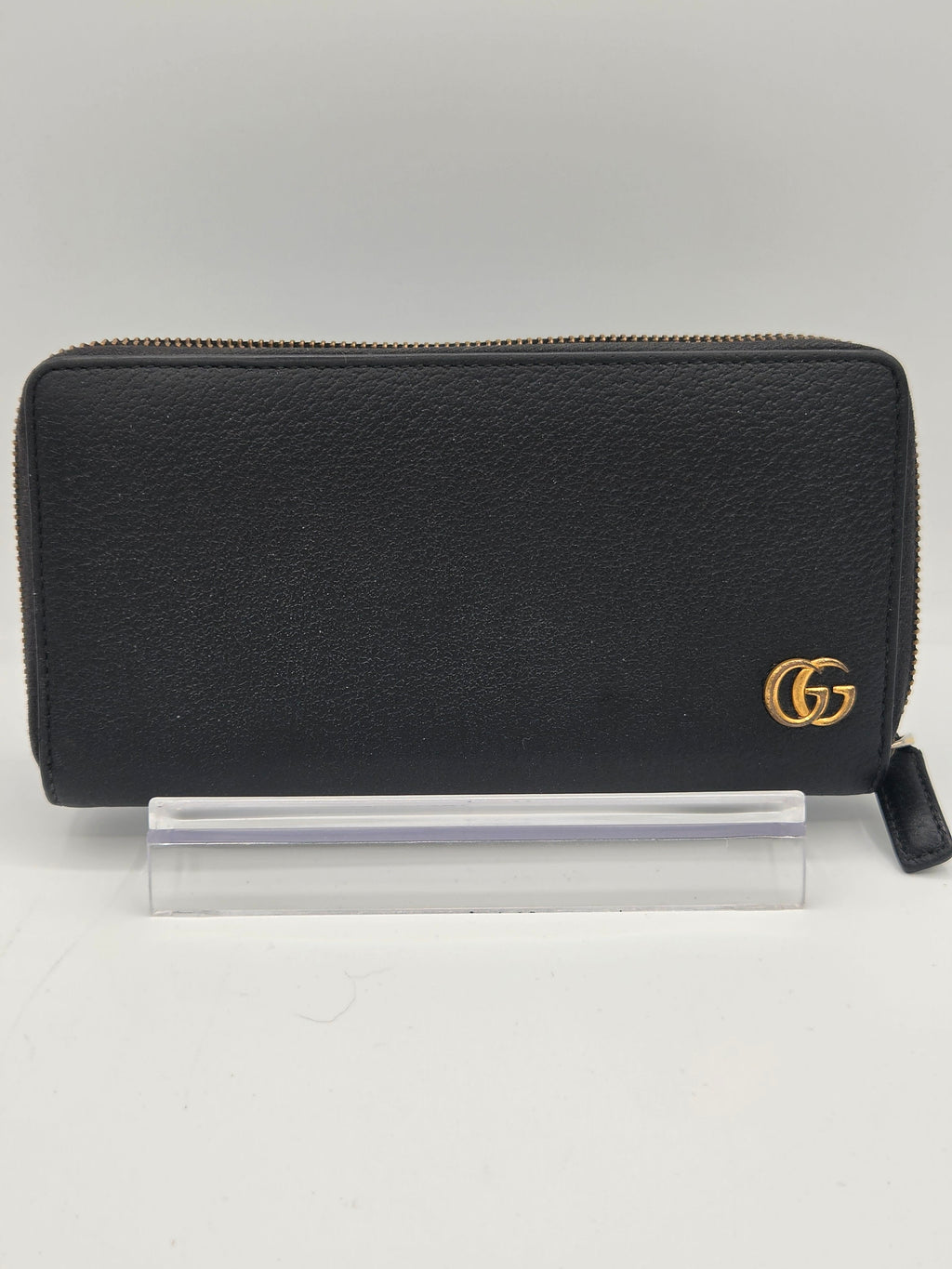 A black leather zip-around wallet with a gold-tone interlocking G logo on the front, shown next to a clear plastic protective cover on a light gray background.