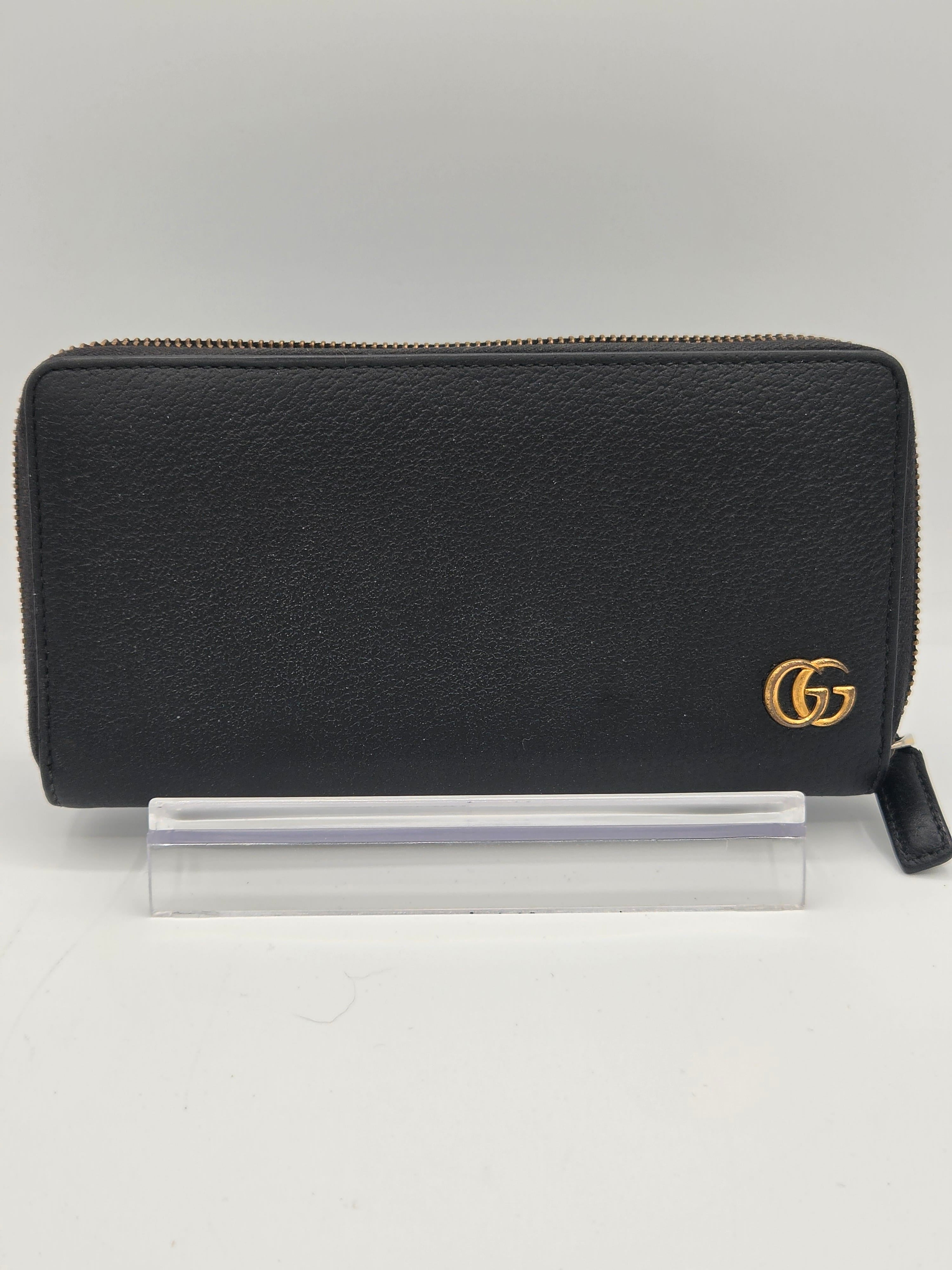 A black leather zip-around wallet with a gold-tone interlocking G logo on the front, shown next to a clear plastic protective cover on a light gray background.