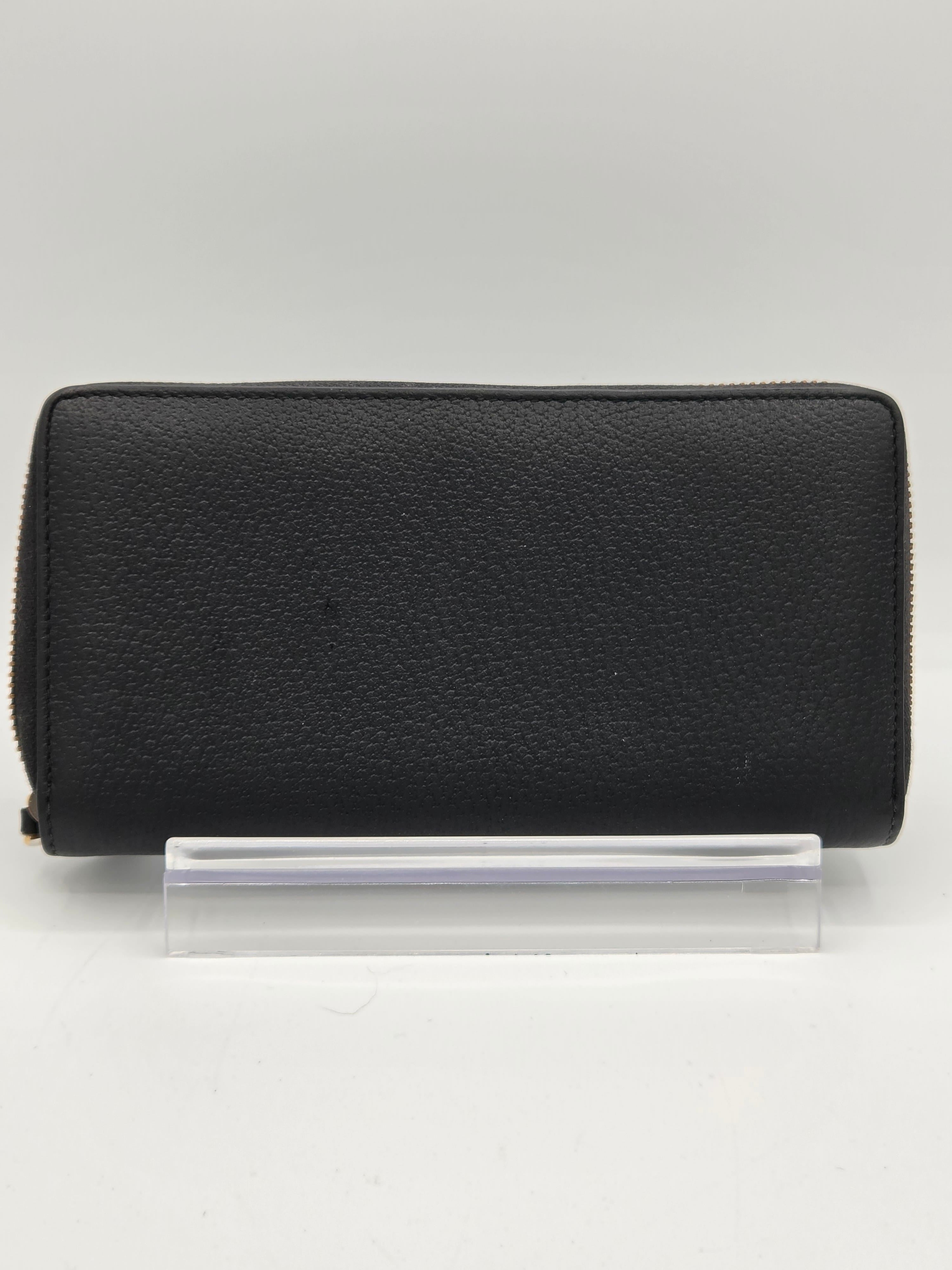 A black pebbled leather zip-around wallet shown closed, positioned next to a clear plastic protective sleeve on a plain white background.