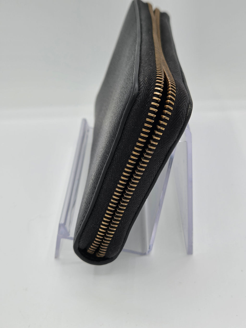 A black leather clutch wallet with a gold-tone zipper, displayed on a clear acrylic stand against a white background.