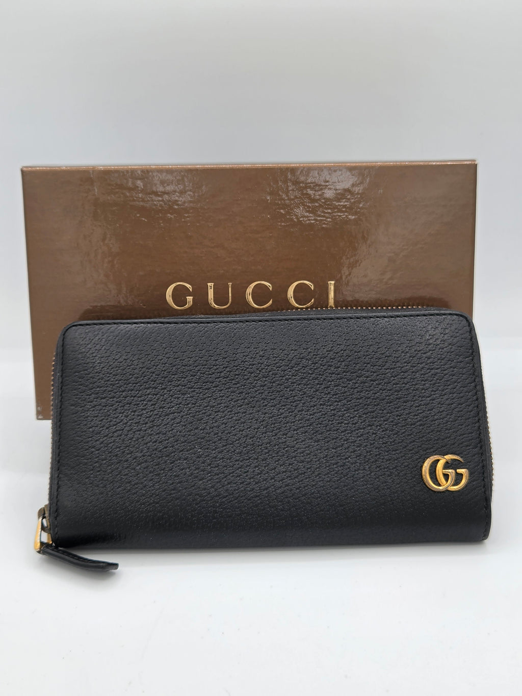 A black pebbled leather Gucci zip-around wallet with a gold-tone interlocking GG logo, displayed next to its brown branded box on a white background.