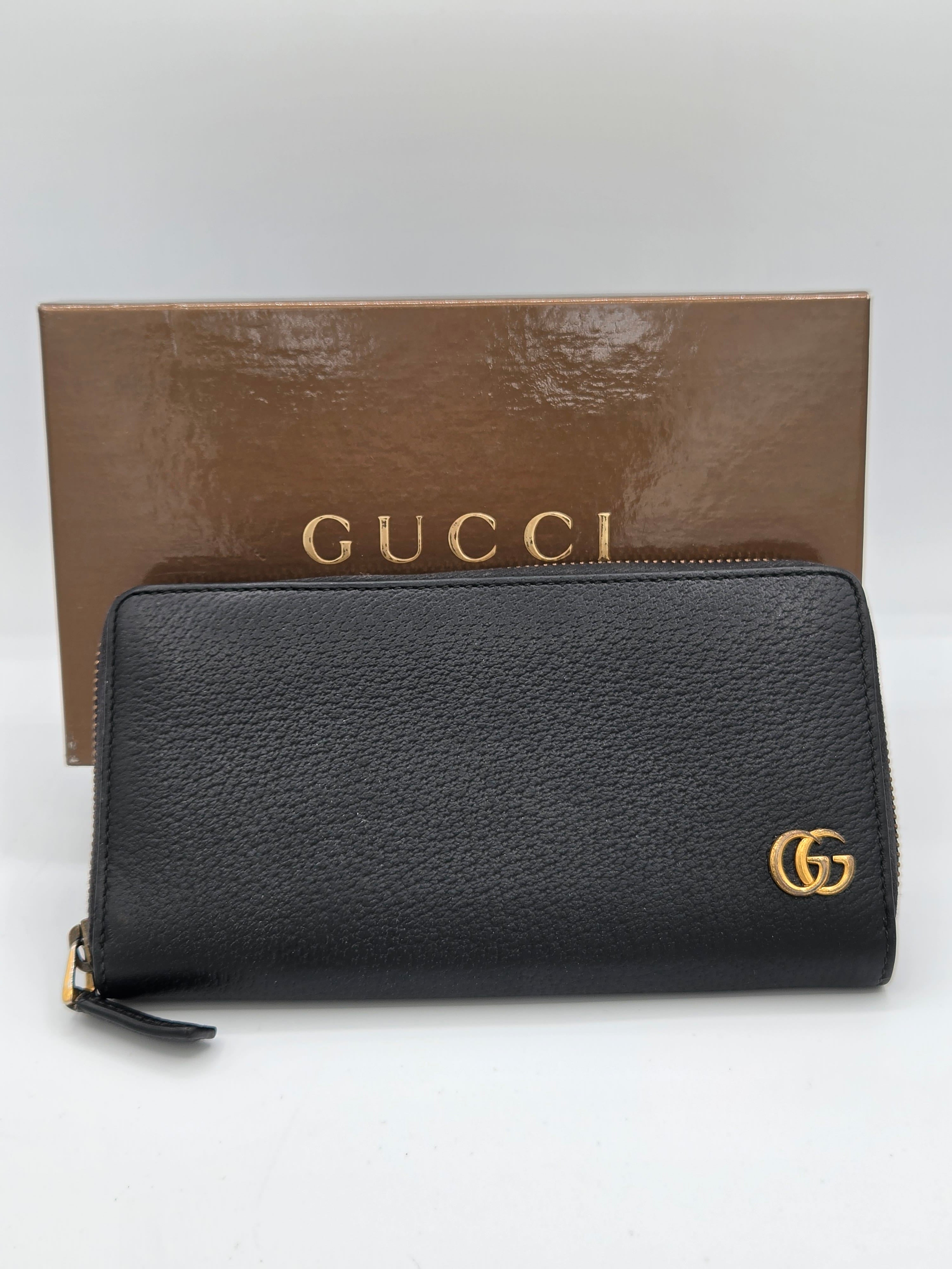 A black pebbled leather Gucci zip-around wallet with a gold-tone interlocking GG logo, displayed next to its brown branded box on a white background.