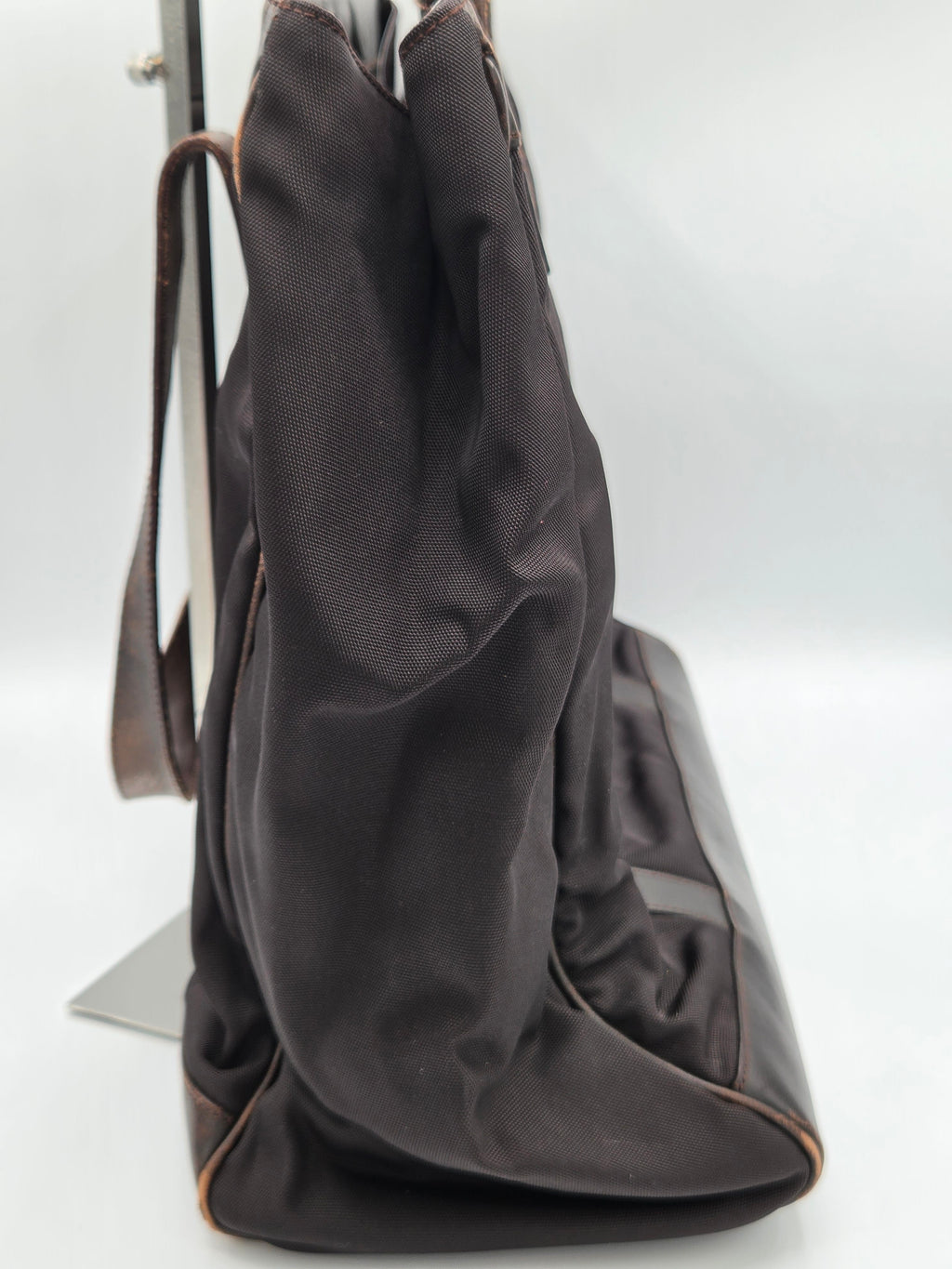 A dark brown fabric shoulder bag with a smooth leather strap, shown resting on a white surface against a white background.
