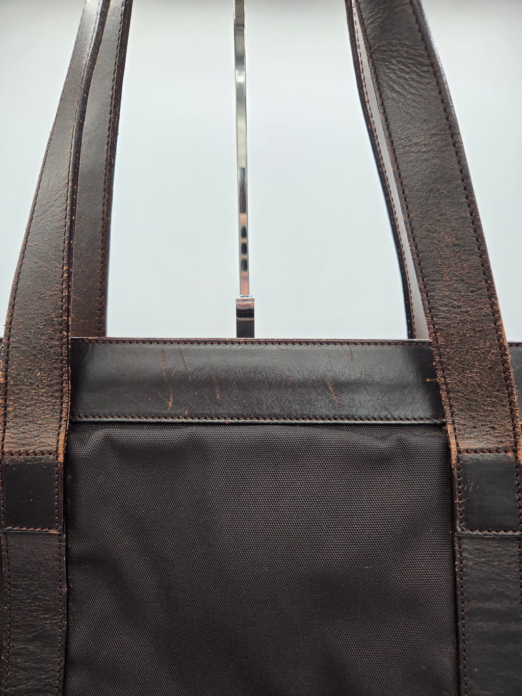 Close-up of a dark brown leather tote bag with a black canvas body, showing the stitching and strap attachments against a light background.