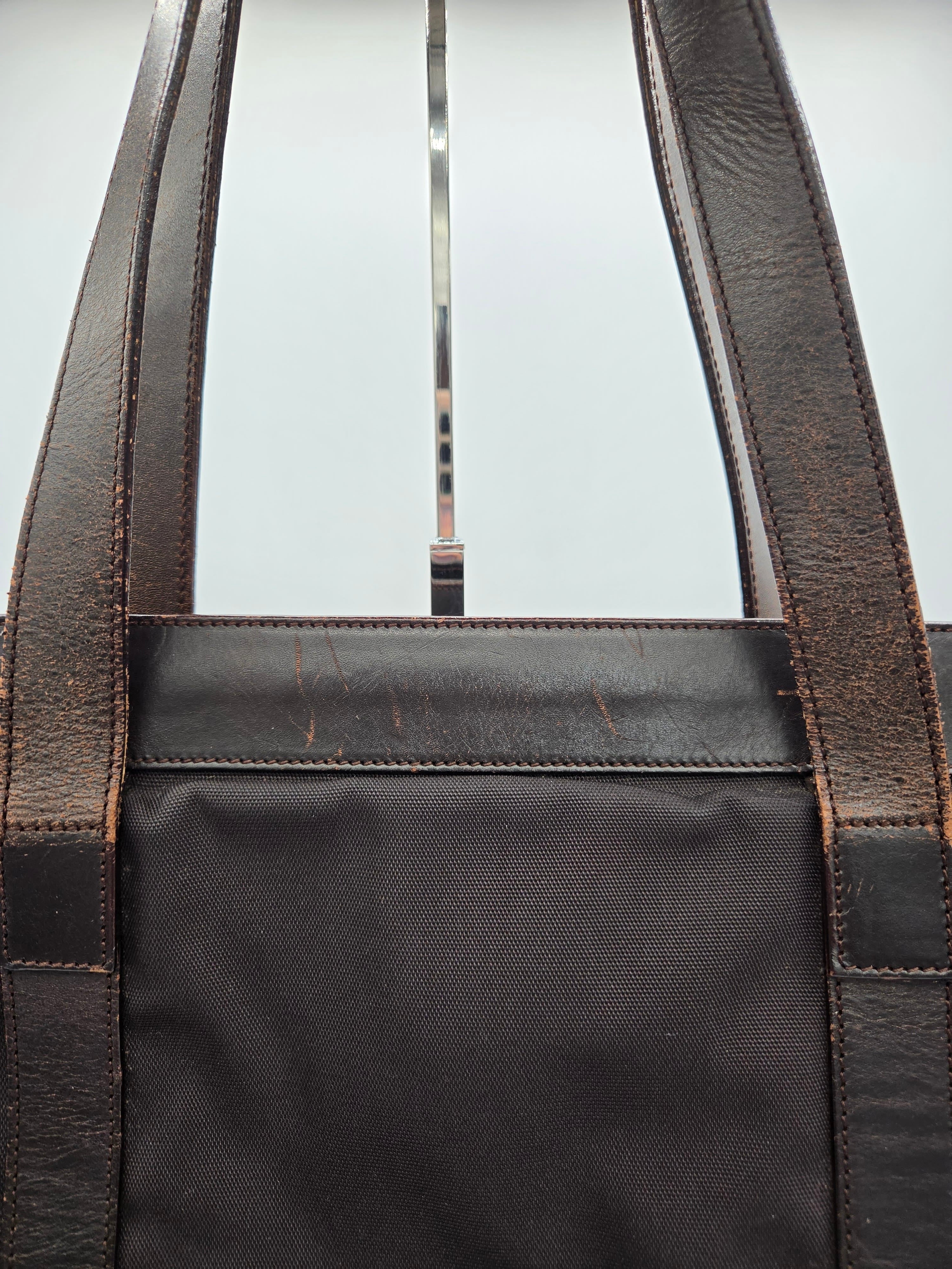 Close-up of a dark brown leather tote bag with a black canvas body, showing the stitching and strap attachments against a light background.