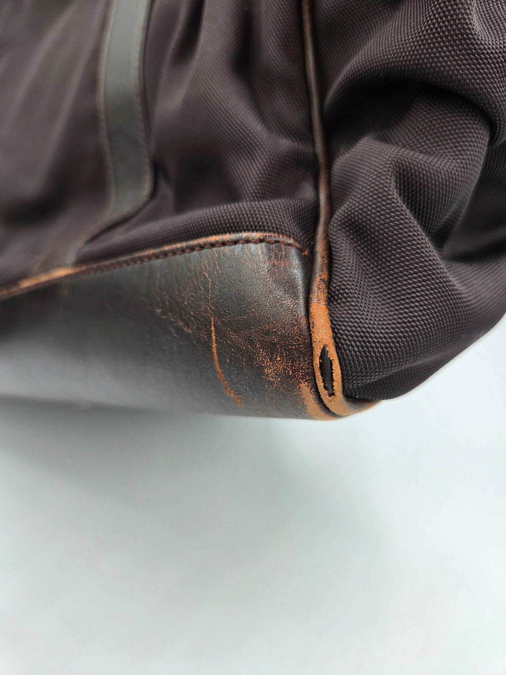 Close-up of a dark brown textured fabric bag corner with a worn, scuffed brown leather trim piece and visible wear marks.