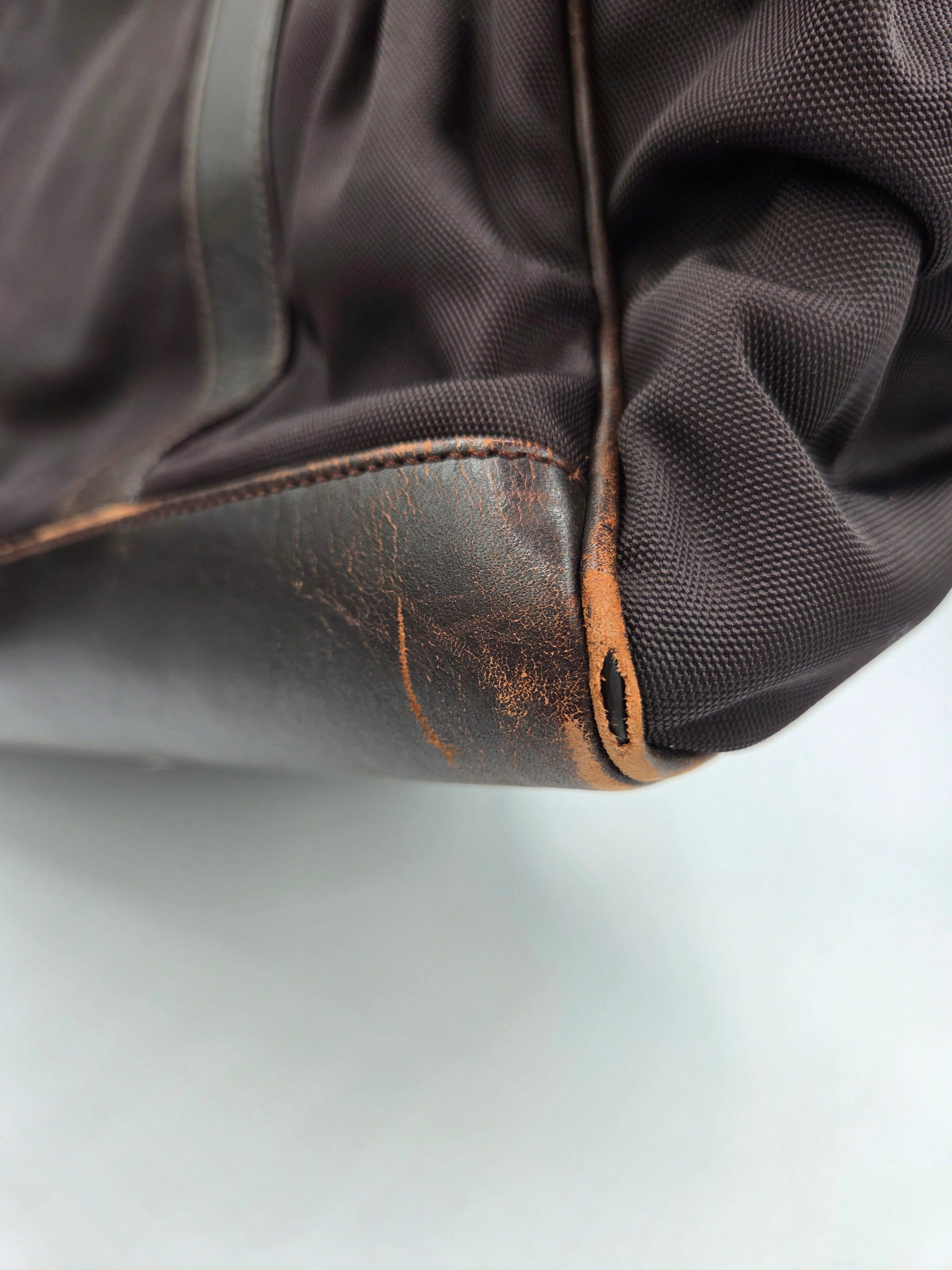 Close-up of a dark brown textured fabric bag corner with a worn, scuffed brown leather trim piece and visible wear marks.
