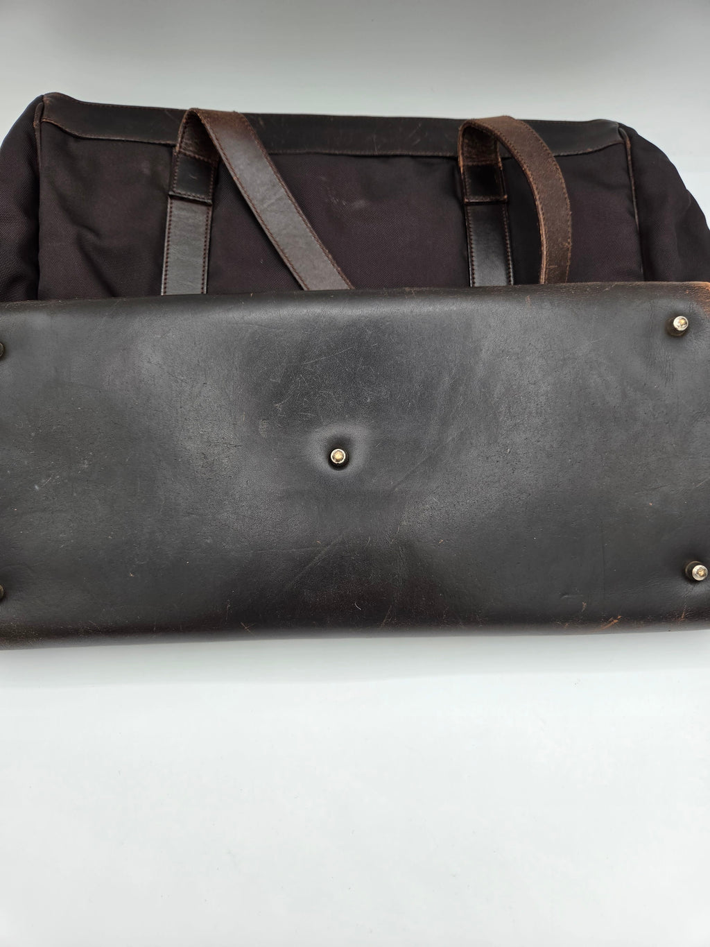 Close-up of a dark brown leather bag or case with metal rivets and straps, positioned next to a black fabric bag on a plain white background.