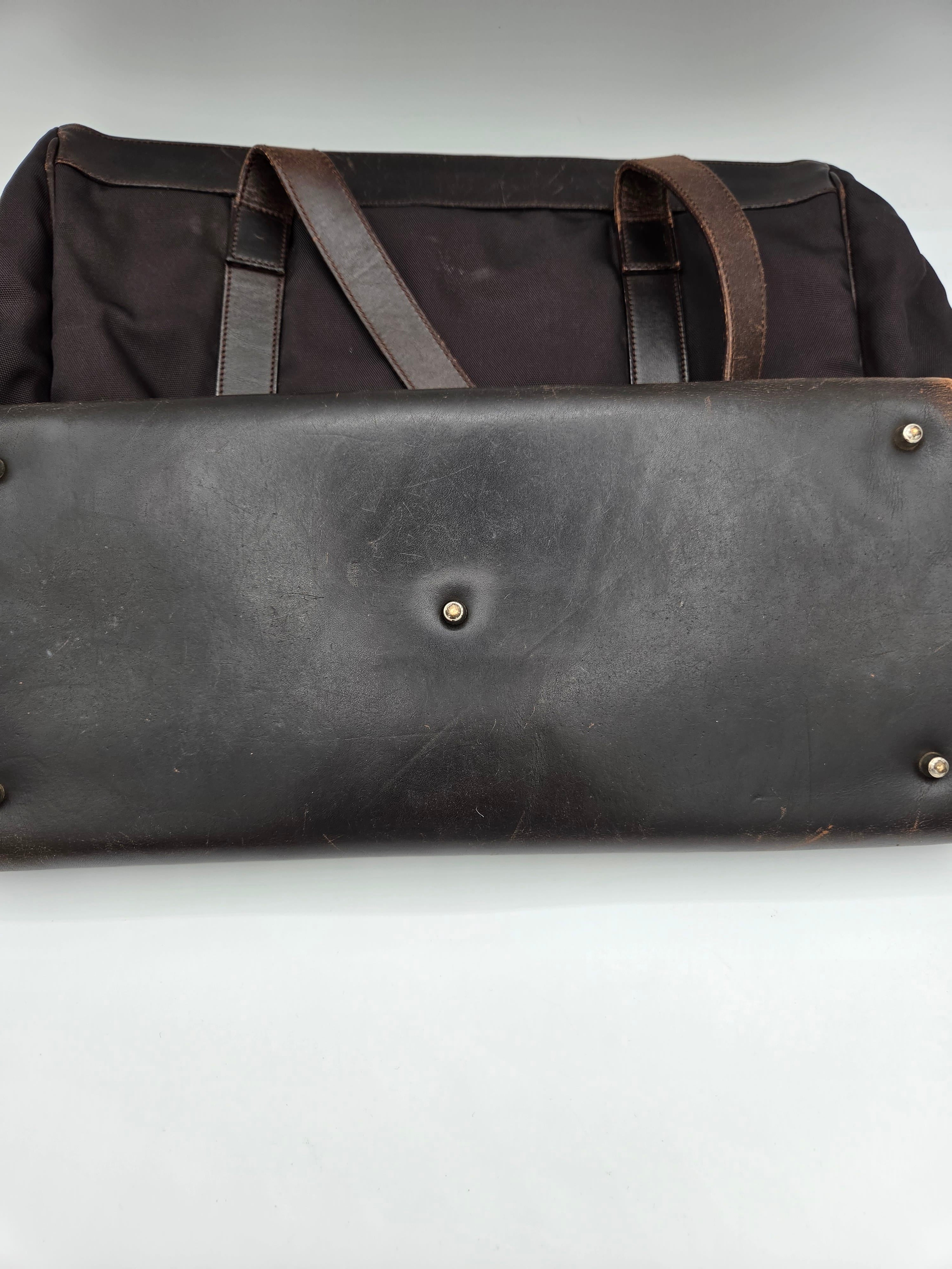 Close-up of a dark brown leather bag or case with metal rivets and straps, positioned next to a black fabric bag on a plain white background.