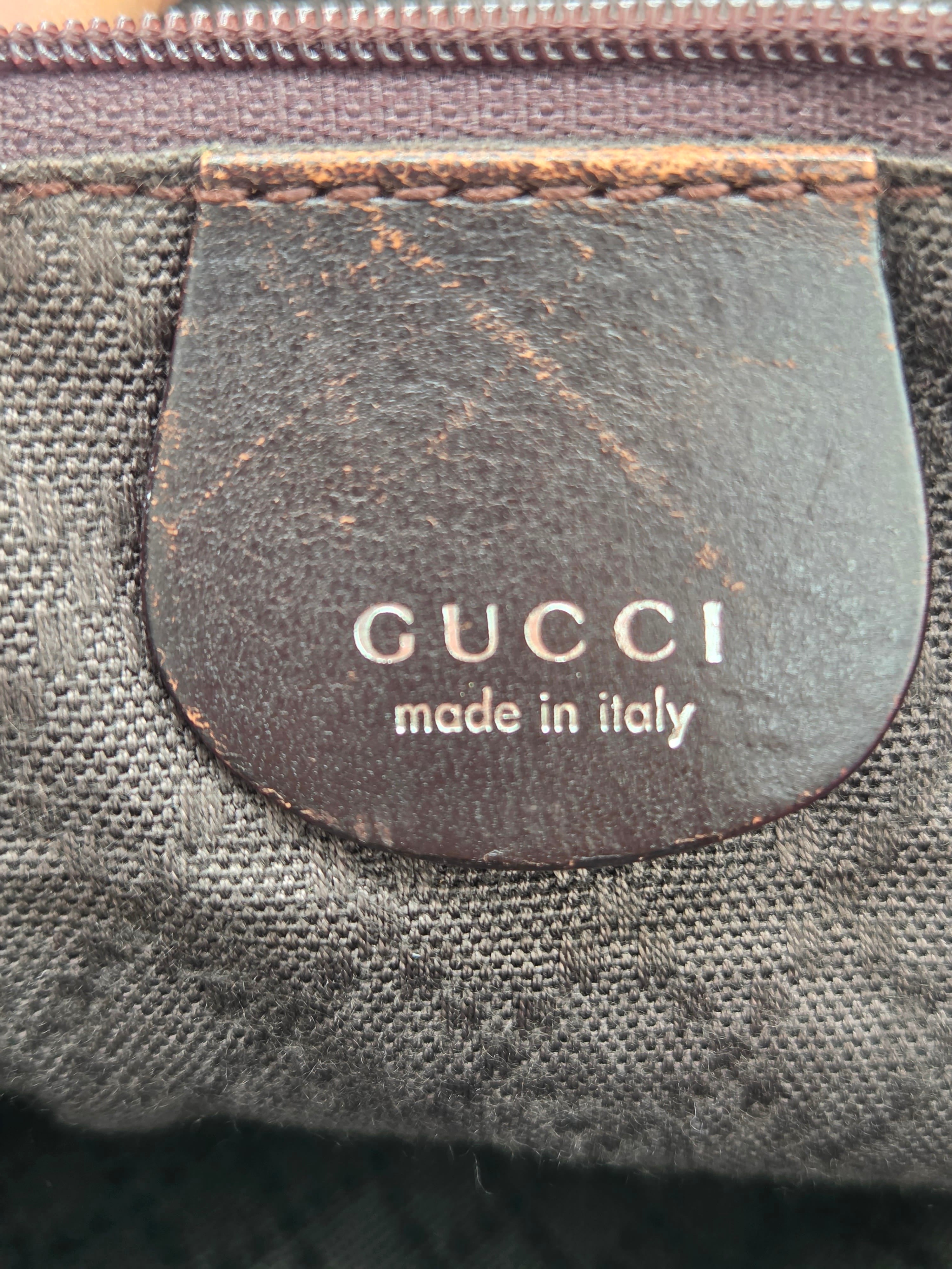 Close-up of a Gucci leather patch with 'GUCCI made in italy' printed in white on a dark brown background, attached to a dark gray textured canvas bag.