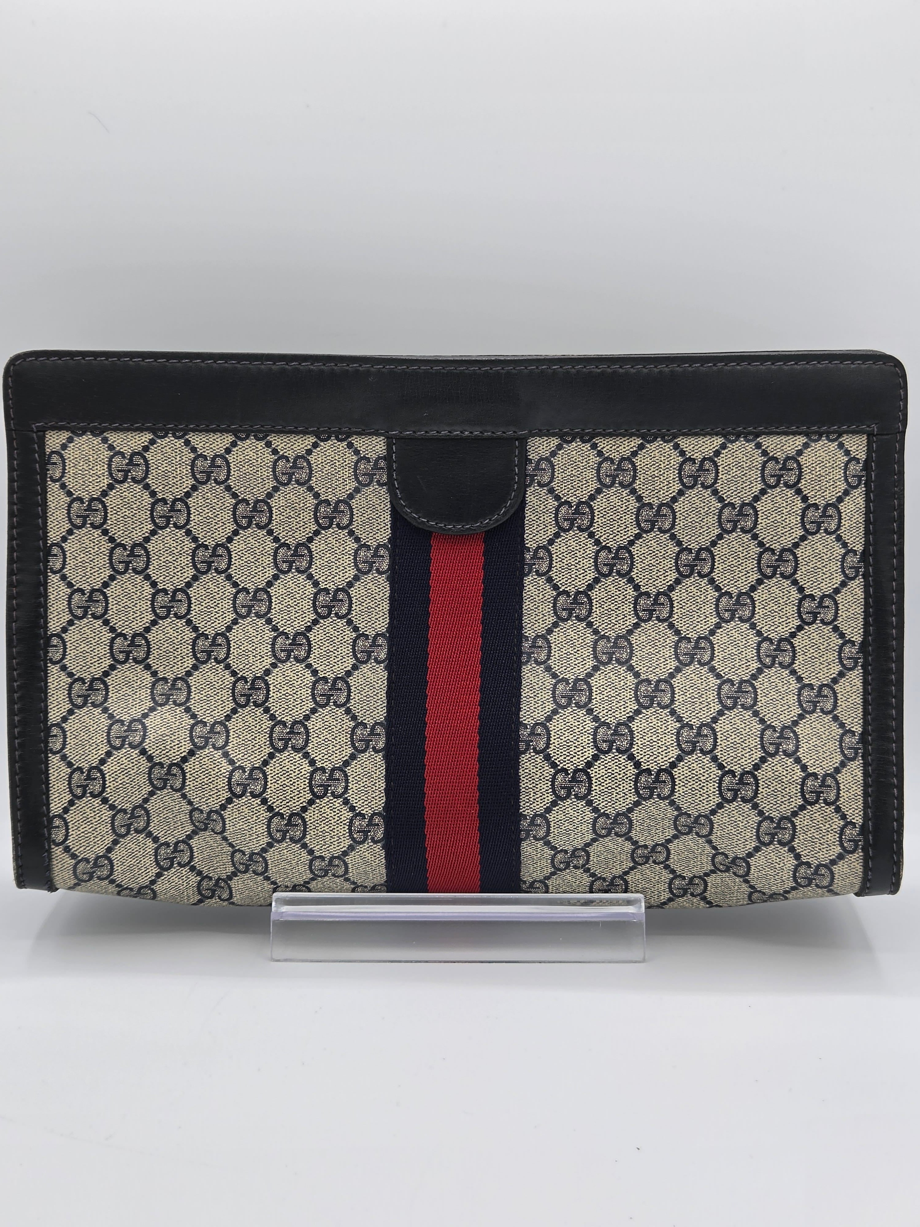 A Gucci monogram canvas document holder or wallet with black leather trim and a signature red and navy green web stripe, shown standing upright against a plain white background.