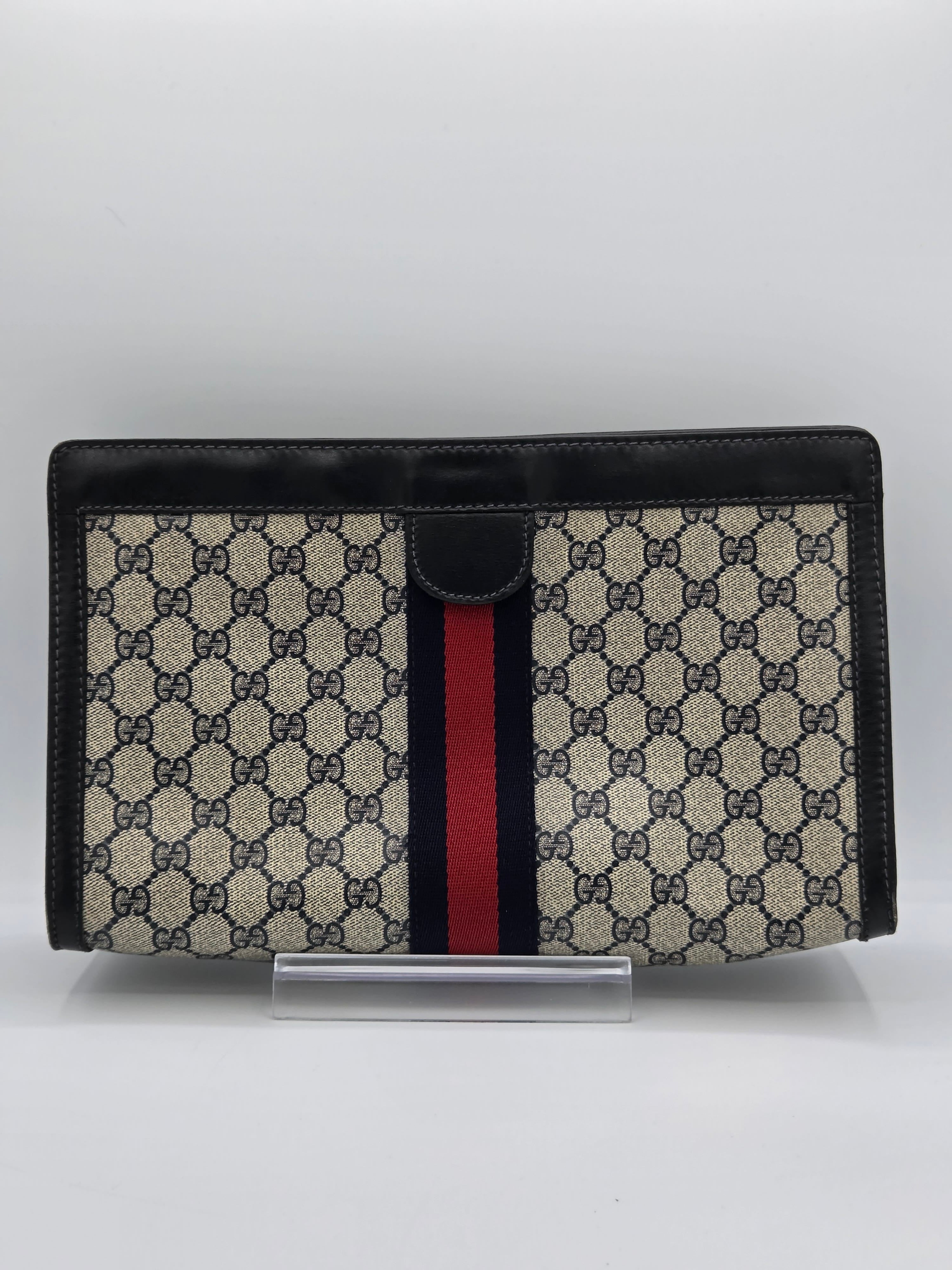 A Gucci monogram canvas document pouch with black leather trim and a signature red and navy green web stripe, shown on a light gray background.