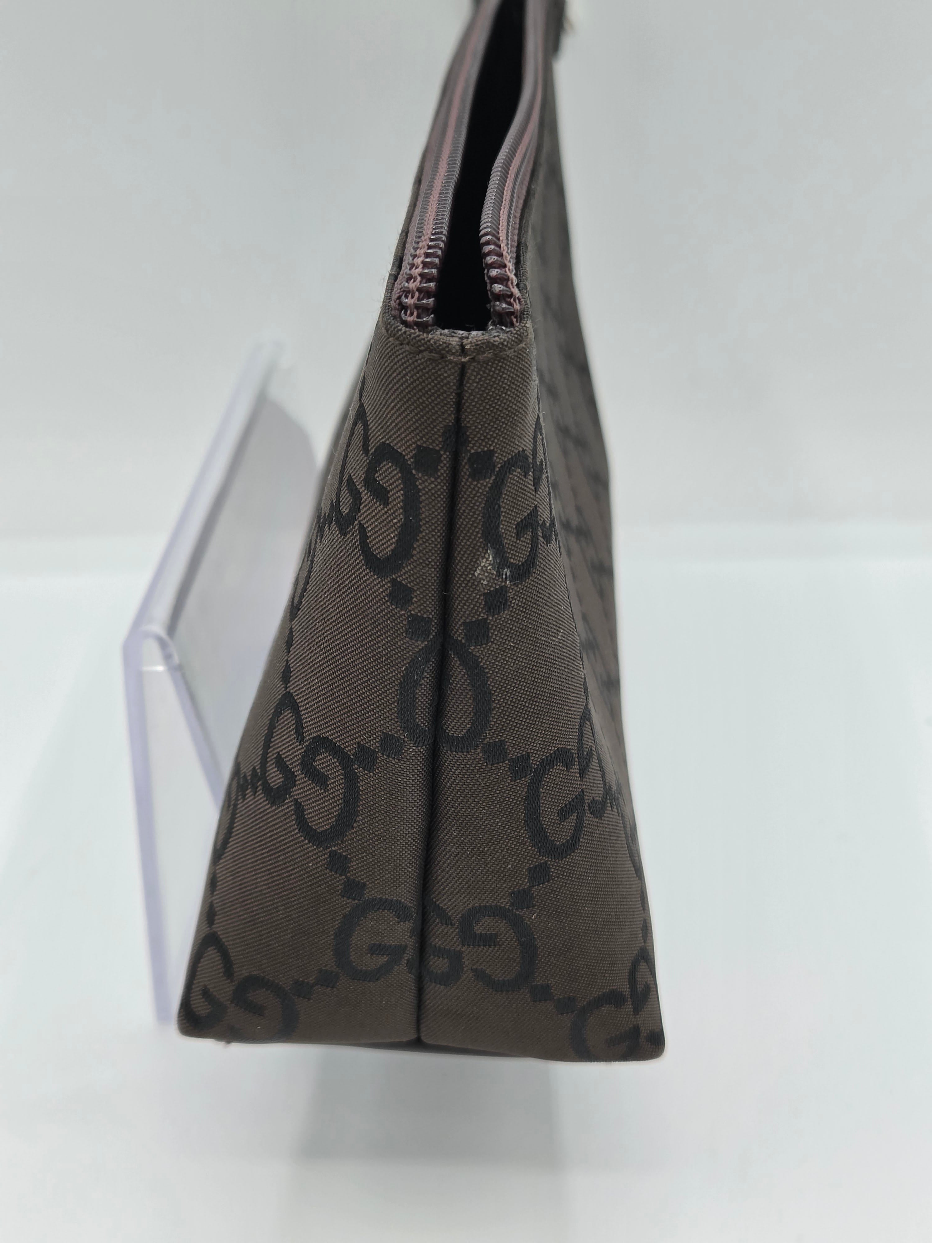 A close-up of a brown monogram pouch or clutch with a repeating interlocking G pattern, shown resting on a clear acrylic display stand against a white background.