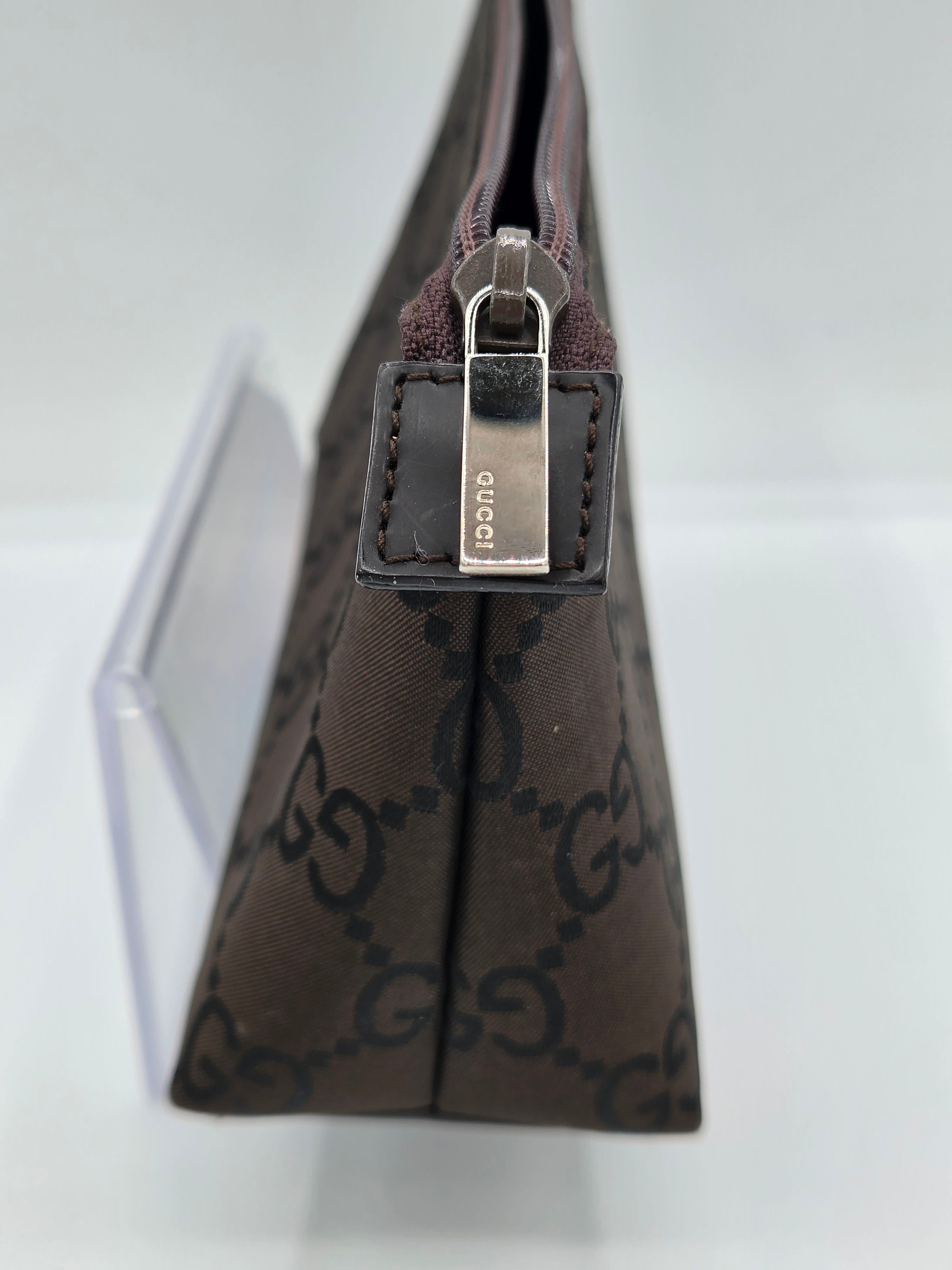 A close-up of a brown Gucci zip pouch featuring the brand's signature GG monogram canvas and a silver-tone metal zipper pull engraved with 'gucci'.