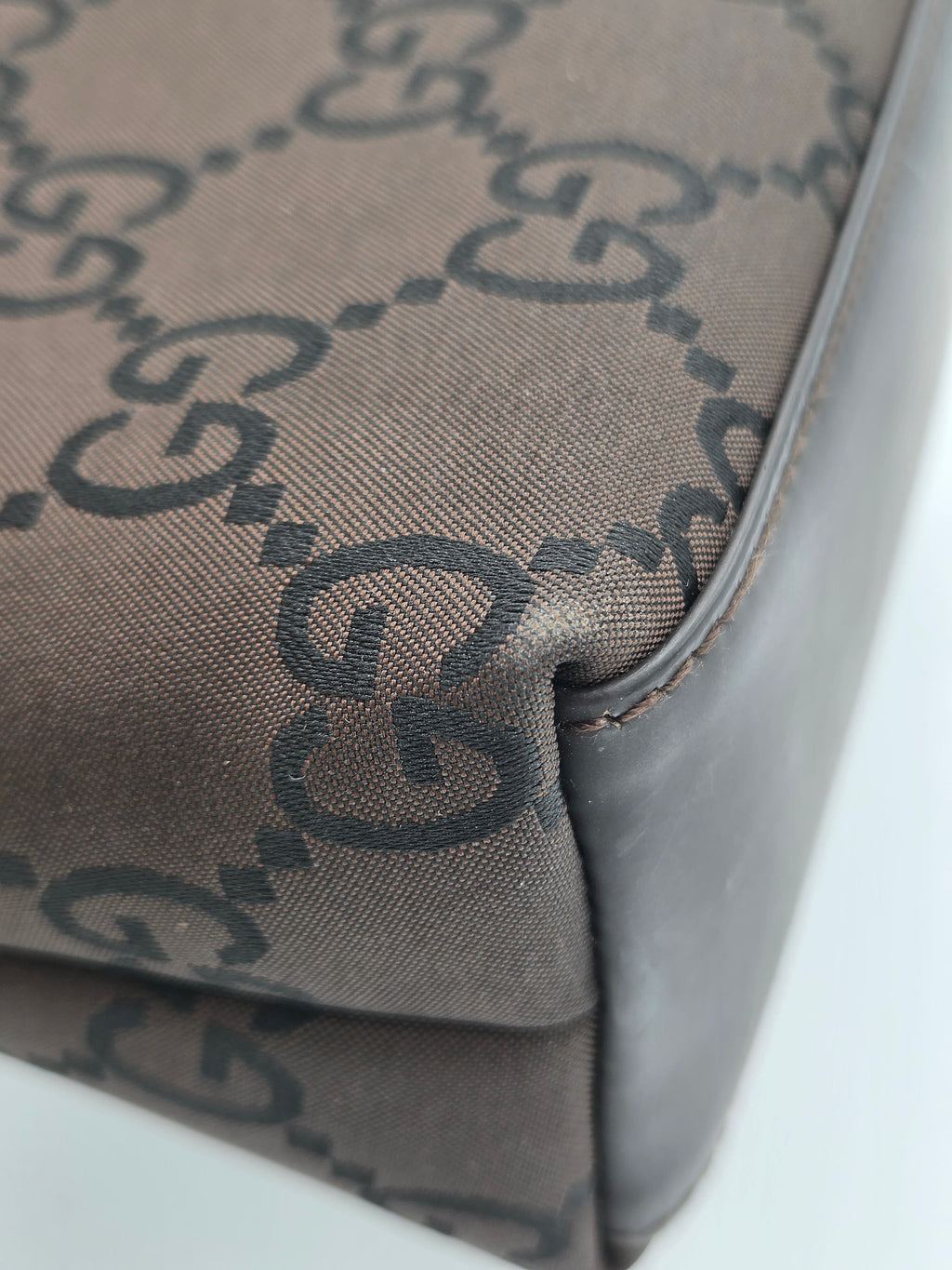 Close-up of a Gucci monogram canvas bag corner with dark brown leather trim and a repeating GG pattern on a taupe background.