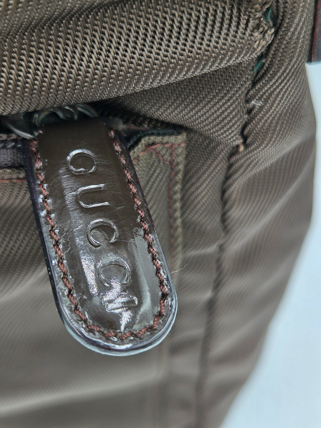 Close-up of a brown woven fabric bag with a dark brown leather zipper pull tab embossed with the brand name 'GUCCI'.