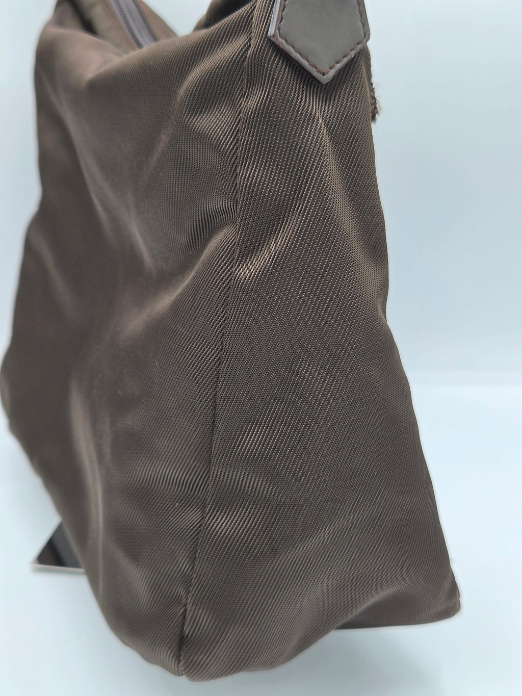 A close-up of a dark brown fabric shoulder bag or satchel, showing the textured woven material and a small dark leather tab on the left side, set against a plain light background.