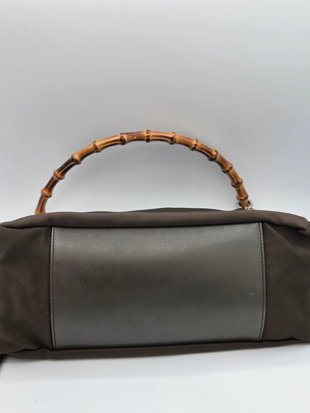 A dark brown fabric handbag with a smooth dark brown leather panel on the front and a distinctive curved bamboo handle, shown against a plain light gray background.