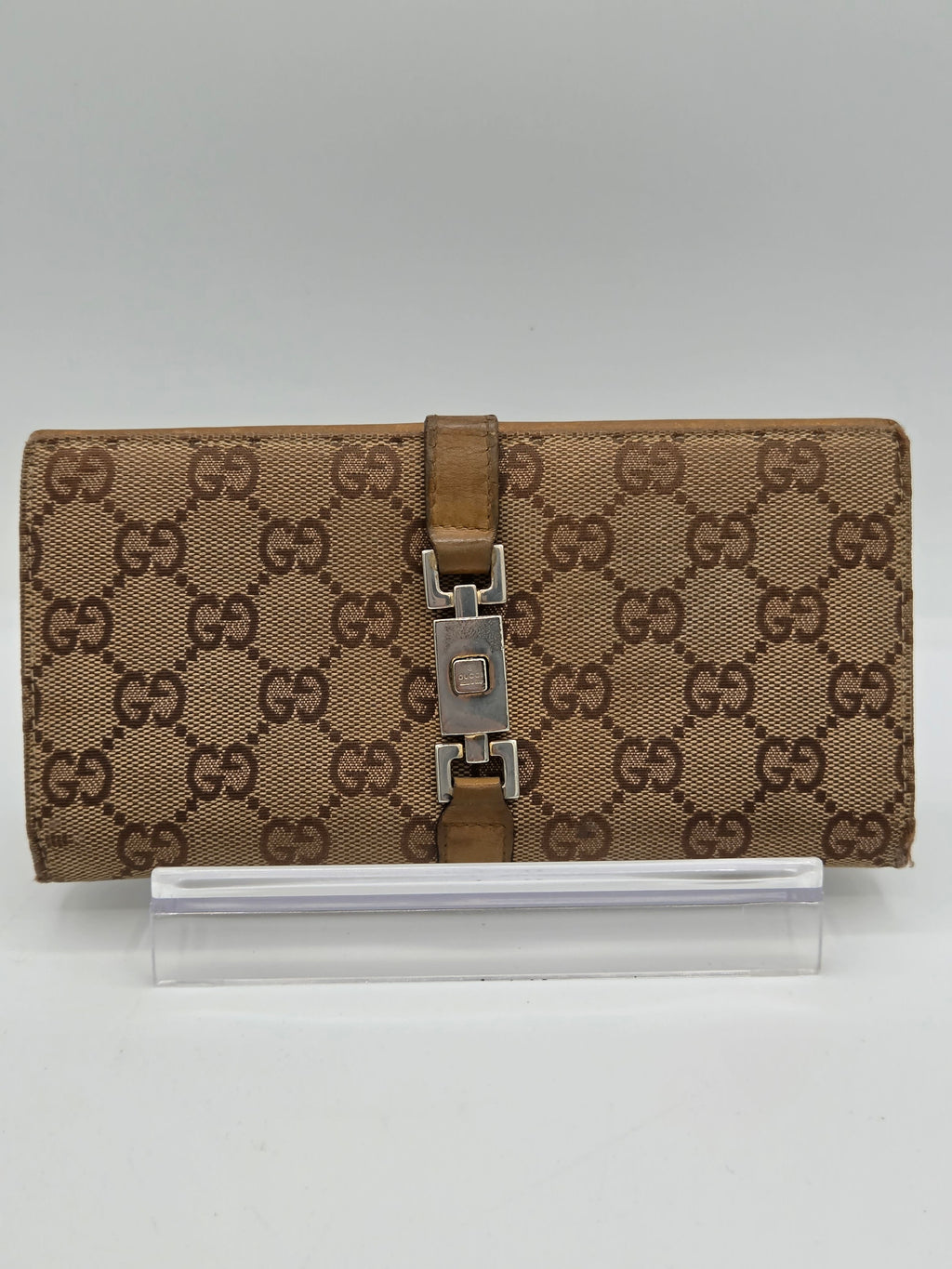 A brown and beige monogram canvas notebook cover with a brown leather strap and silver metal buckle, shown next to a clear plastic sleeve on a light gray background.