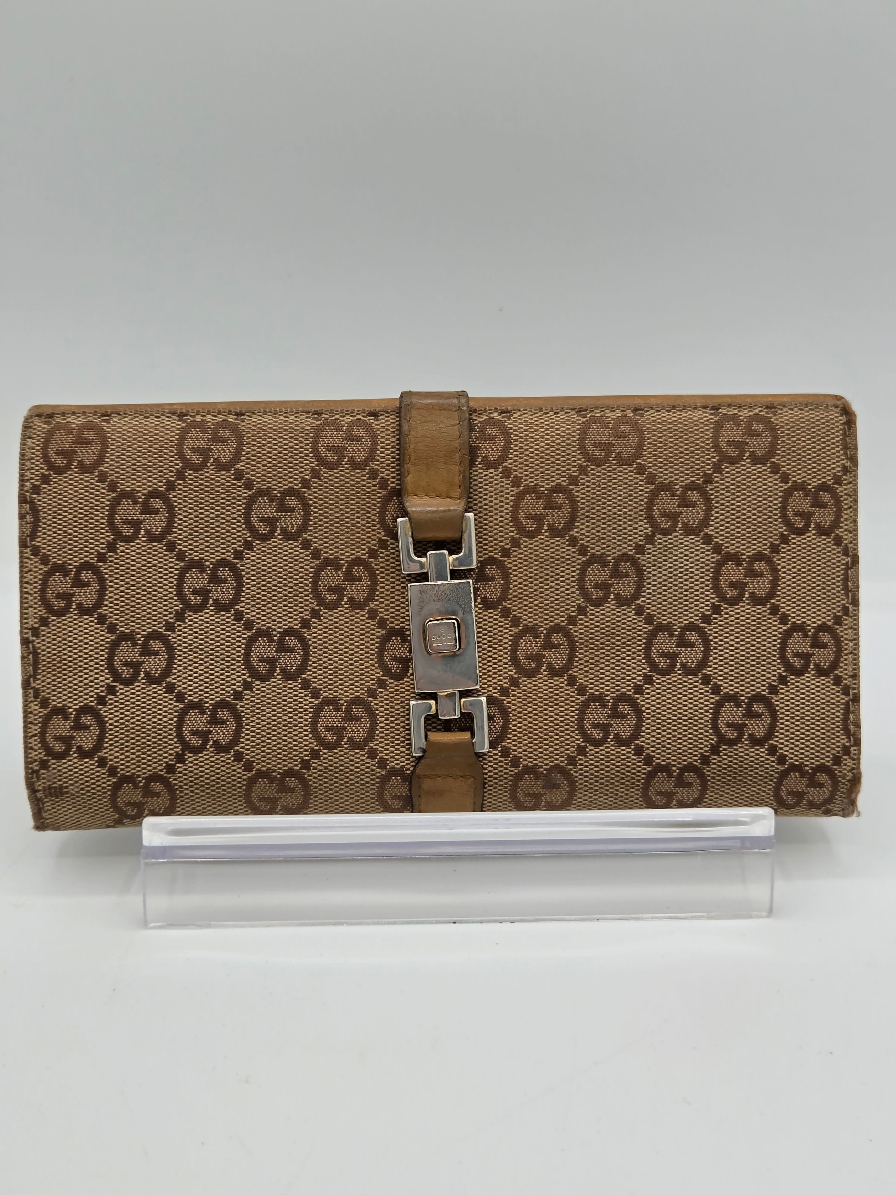 A brown and beige monogram canvas notebook cover with a brown leather strap and silver metal buckle, shown next to a clear plastic sleeve on a light gray background.
