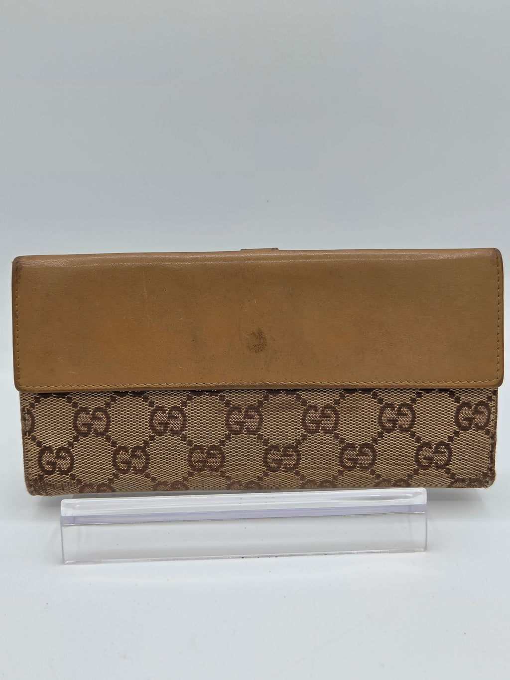 A vintage Gucci wallet or clutch in brown GG Supreme canvas and tan leather, shown next to a clear plastic sleeve on a light gray background.